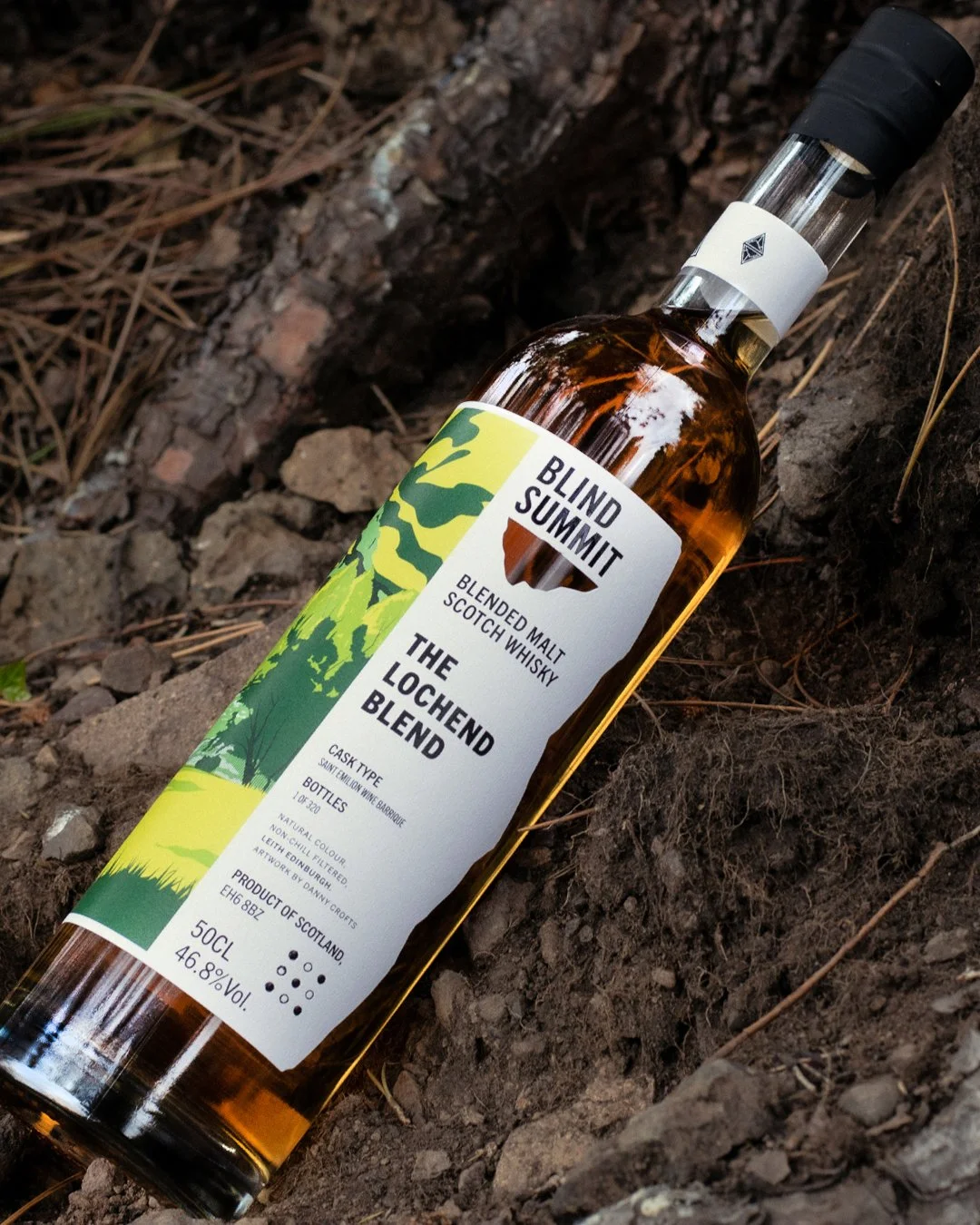 A bottle of blended malt Scotch whisky called 'The Lochend Blend' lying on the ground among dirt and pine needles in a natural outdoor setting.