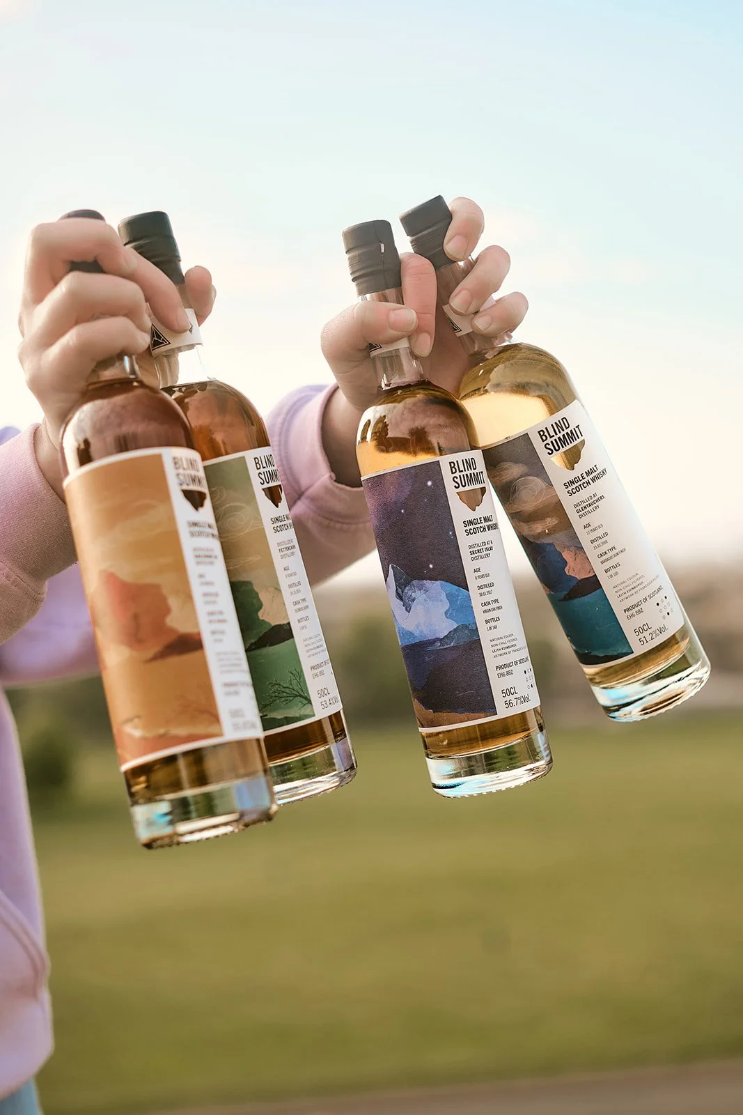 A woman holding bottles of whisky with custom labels outdoors, sky and blurred landscape background.
