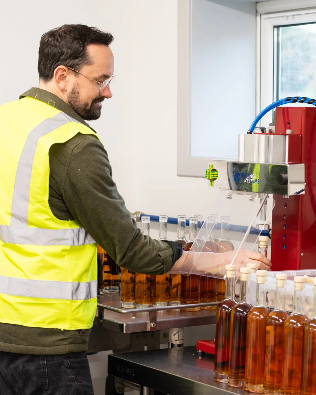 A man wearing safety glasses, a yellow safety vest, and dark clothing is working with a filling machine, filling bottles with amber-colored liquid inside a lab or production facility.