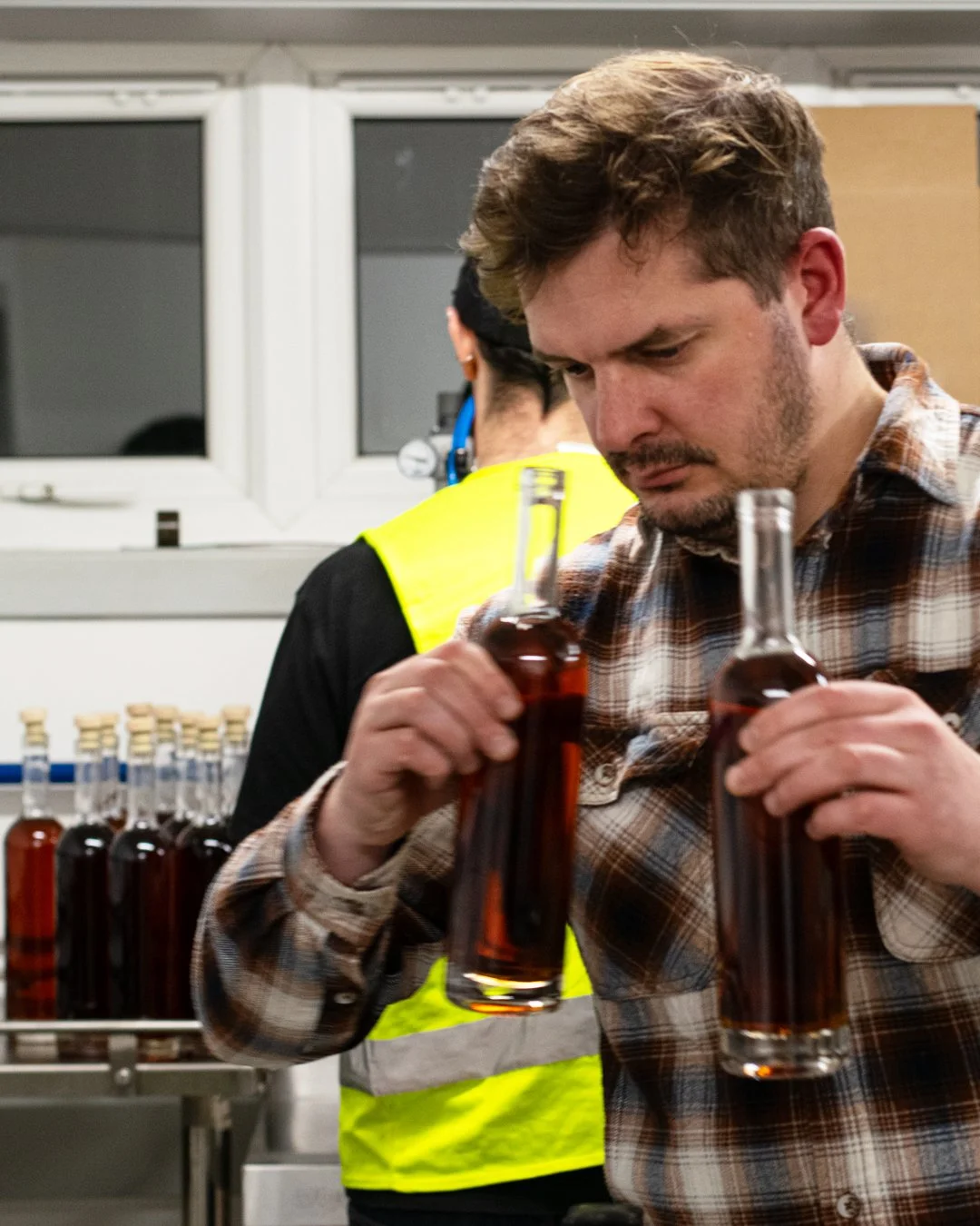 A man wearing a plaid shirt examining two glass bottles filled with dark liquid in a factory or production setting, with a person in a high-visibility vest working in the background.
