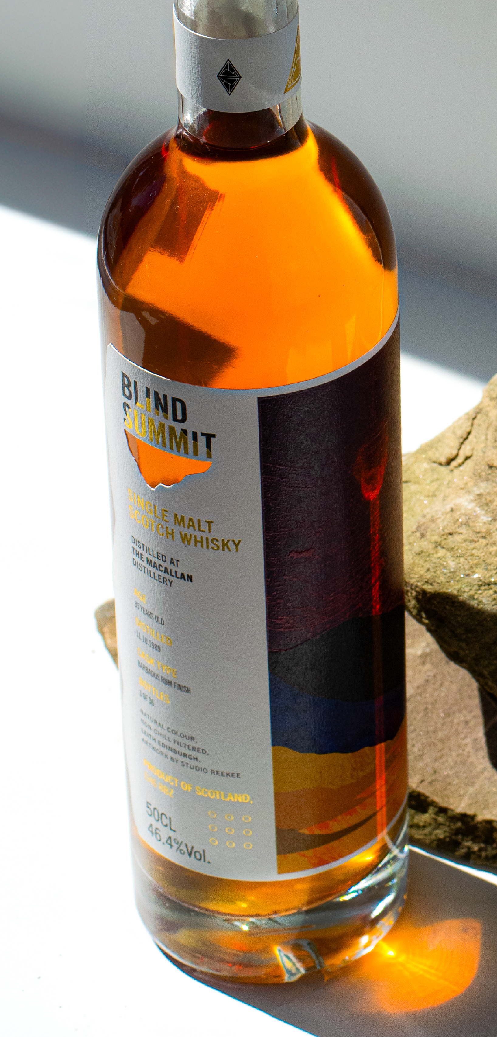 A bottle of Blind Summit Macallan, displayed on a rock with sunlight reflecting through the amber liquid.