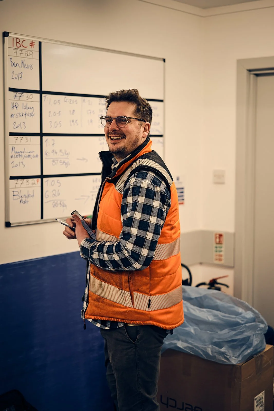 A smiling man with glasses, wearing an orange safety vest over a plaid shirt, standing in front of a whiteboard with handwritten notes.
