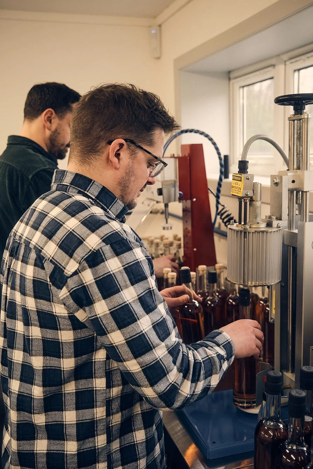Two men working in a laboratory with glass bottles, machinery, and equipment, possibly filling or inspecting the bottles.