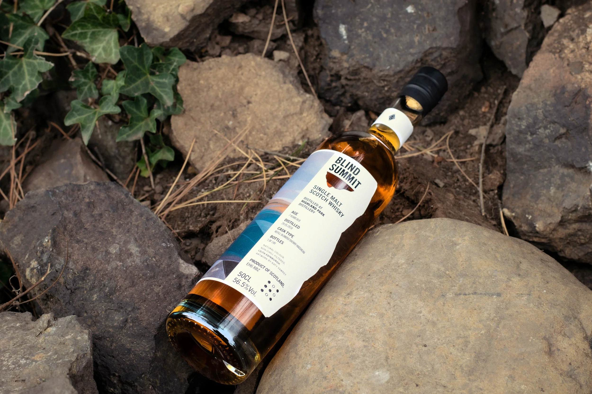 A bottle of Blind Summit single malt Scotch whisky lying on rocks surrounded by dirt and some green leaves.