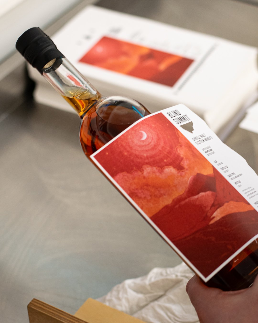 A hand holding a bottle of whiskey with a red and orange sunset mountain landscape label, in front of a document or brochure with similar artwork.