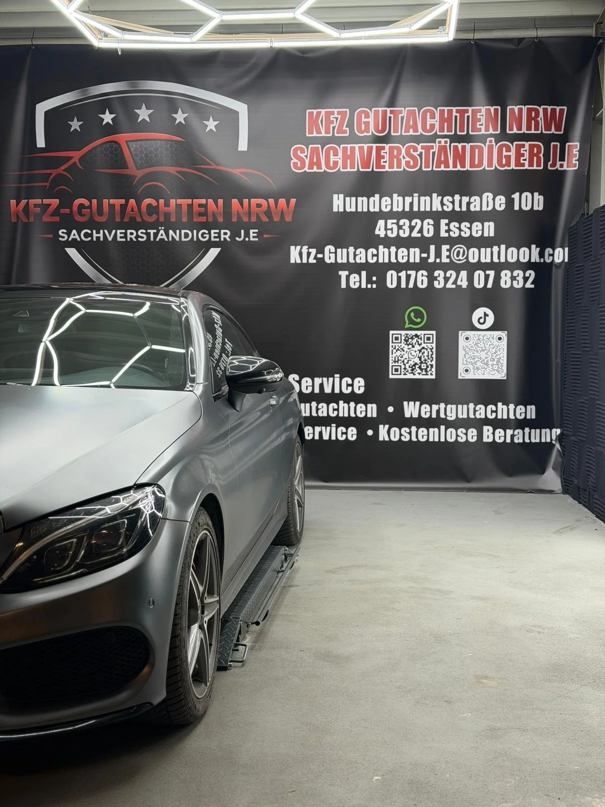 Ein schwarzer Hintergrund mit einem roten und weißen Schriftzug, eine Logo mit einem stilisierten Auto, und ein graues Auto auf einer Werkstattbock. Es sind Kontaktdaten und Hinweise auf Dienstleistungen wie Gutachten, Service und kostenlose Beratung.