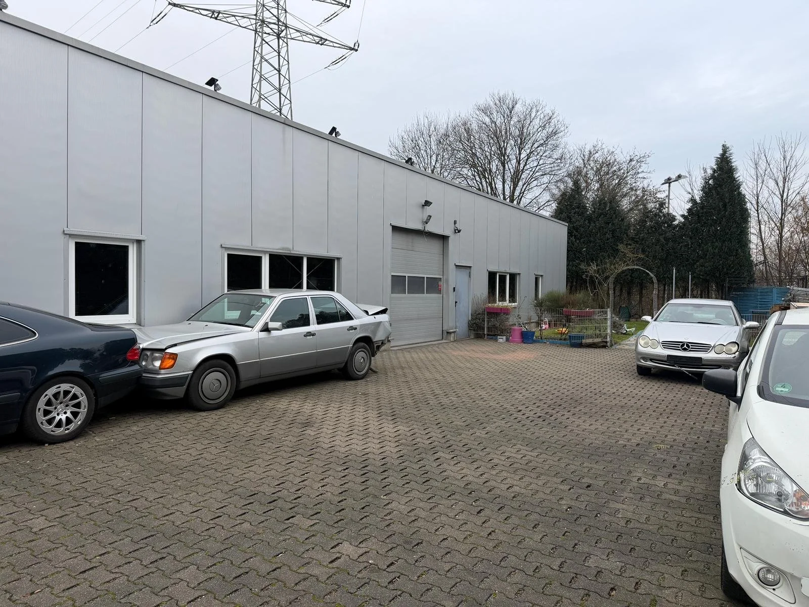 Ein Parkplatz vor einer Industriehalle mit mehreren geparkten Autos und einem kleinen Gartenbereich mit Spielzeug und Pflanzen.