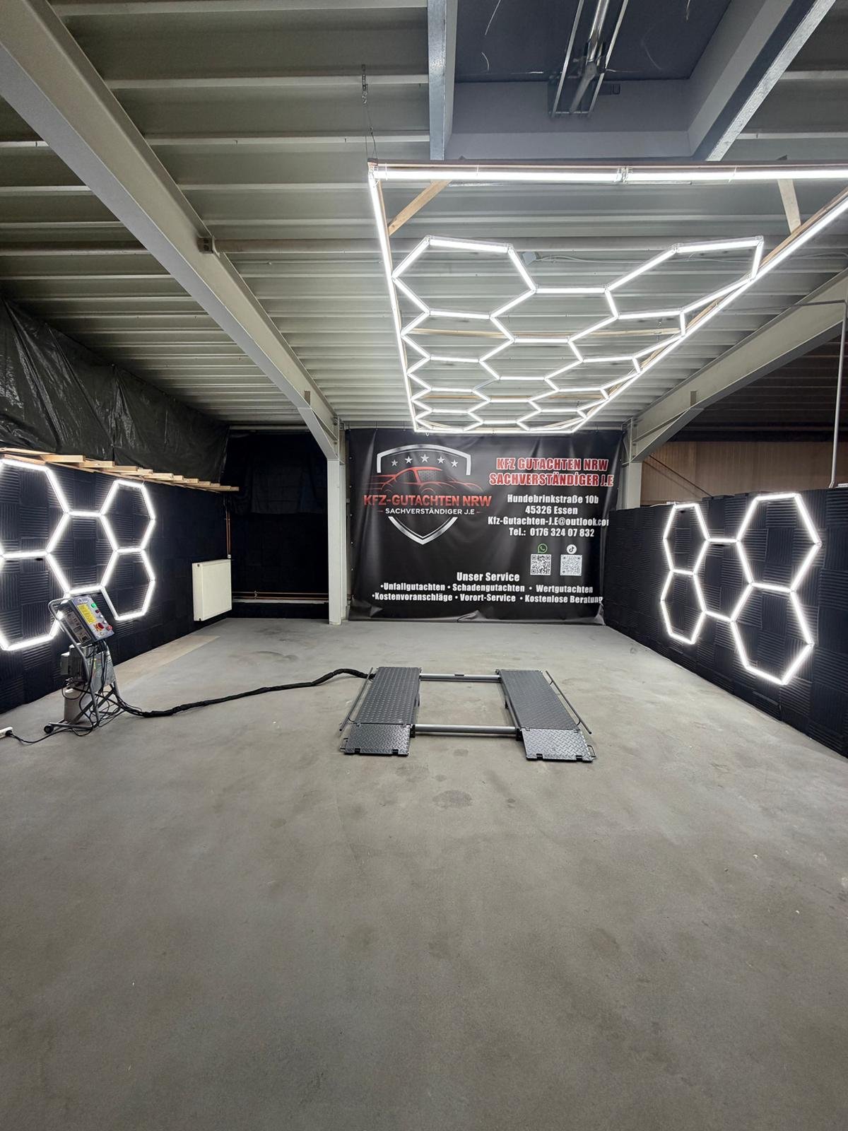 Ein leerer Raum mit Betonboden, an den Wänden sind LED-Leuchten in Form von Hexagonen angebracht. Im Hintergrund befindet sich ein schwarzer Vorhang mit einem Logo und deutschen Text, der auf eine KFZ-Gutachtenfirma hinweist. In der Mitte des Raumes steht eine Fahrzeugwaage, und auf der linken Seite ist eine Reinigungsmaschine.
