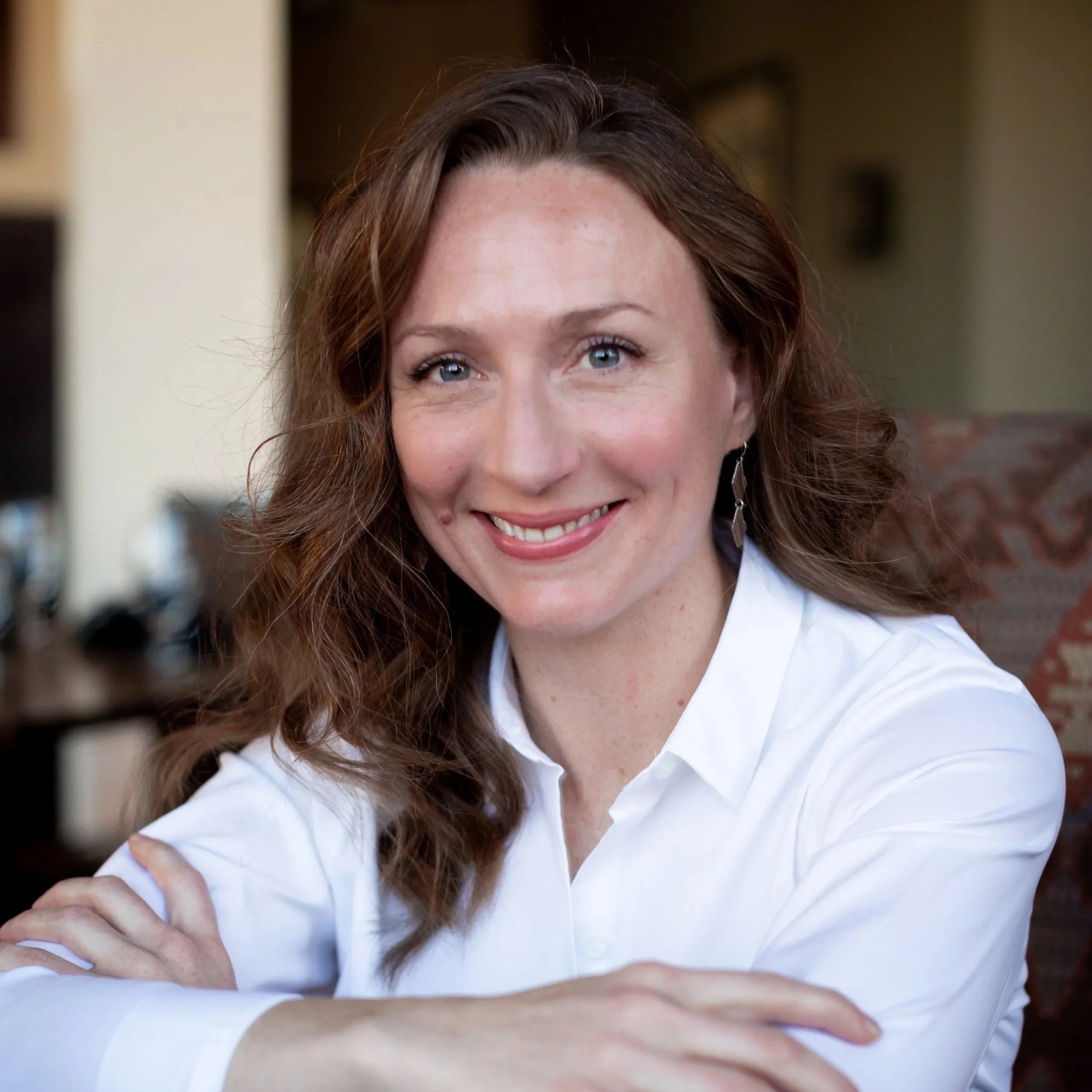 A woman with wavy brown hair, blue eyes, and fair skin smiling and wearing a white button-up shirt, sitting indoors.