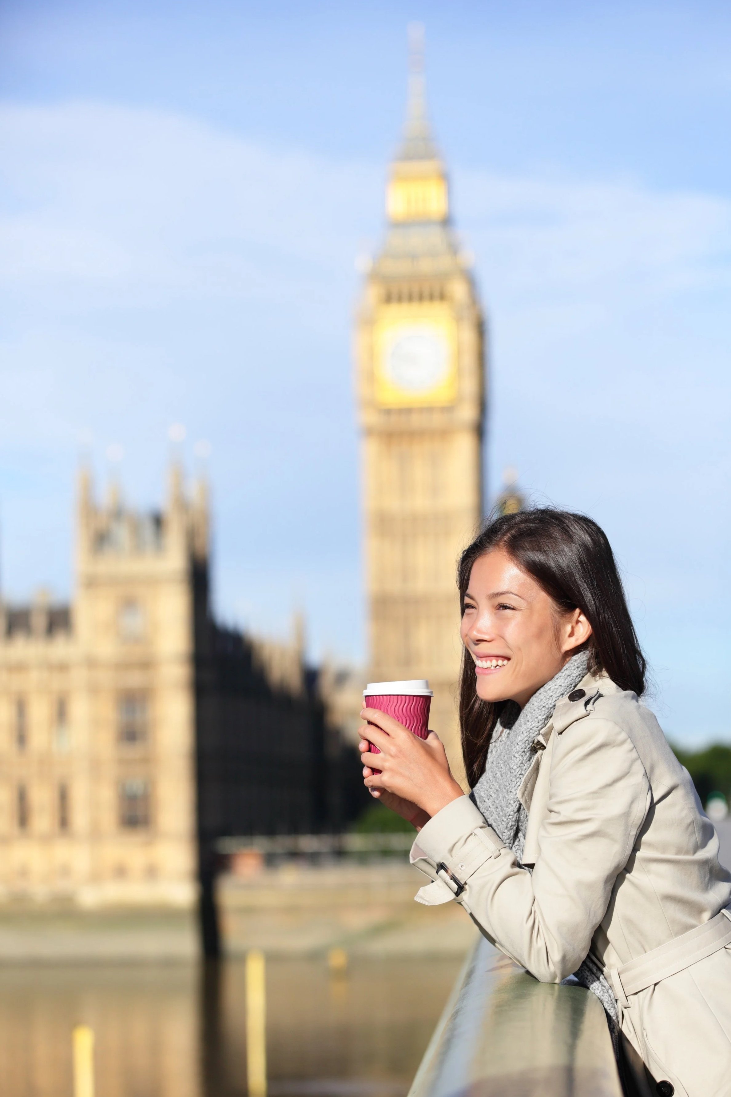 A woman with dark hair smiling and holding a pink coffee cup outdoors with Big Ben and the Houses of Parliament in London in the background.