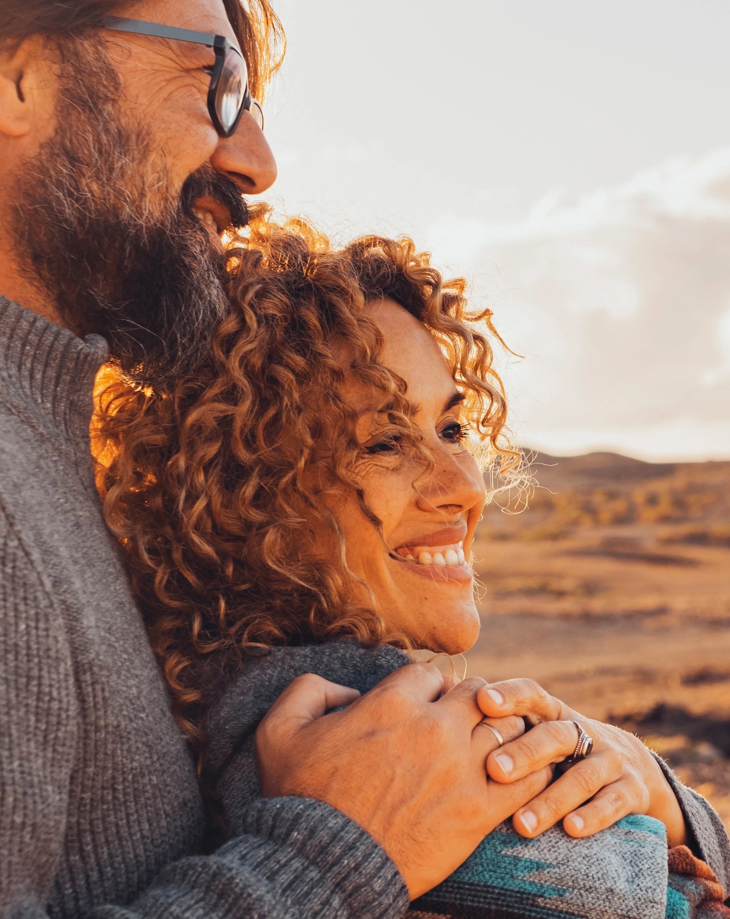 A smiling couple with curly hair embracing in an outdoor landscape at sunset.