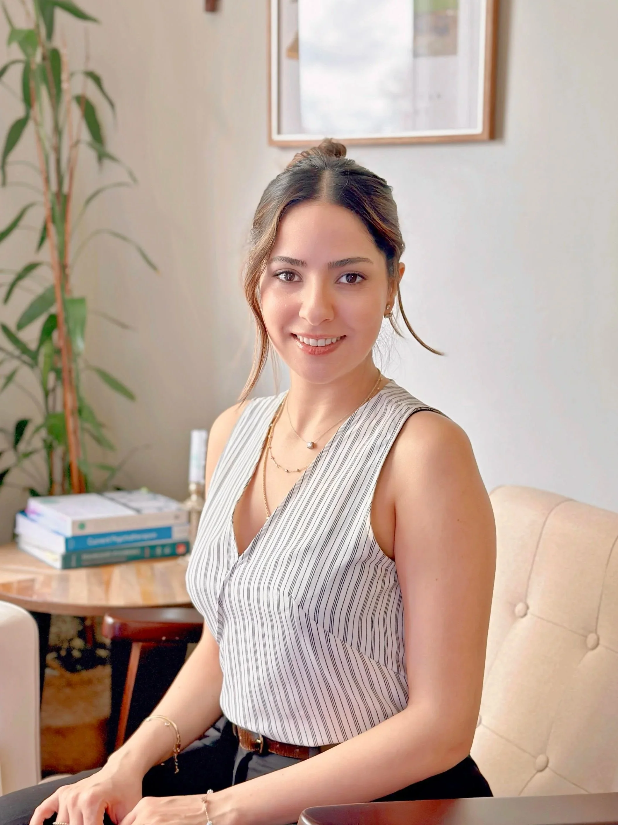 A young woman with brown hair tied up, smiling, sitting on a beige chair in an office. She is wearing a sleeveless, striped blouse and jewelry. There are books and a plant in the background.