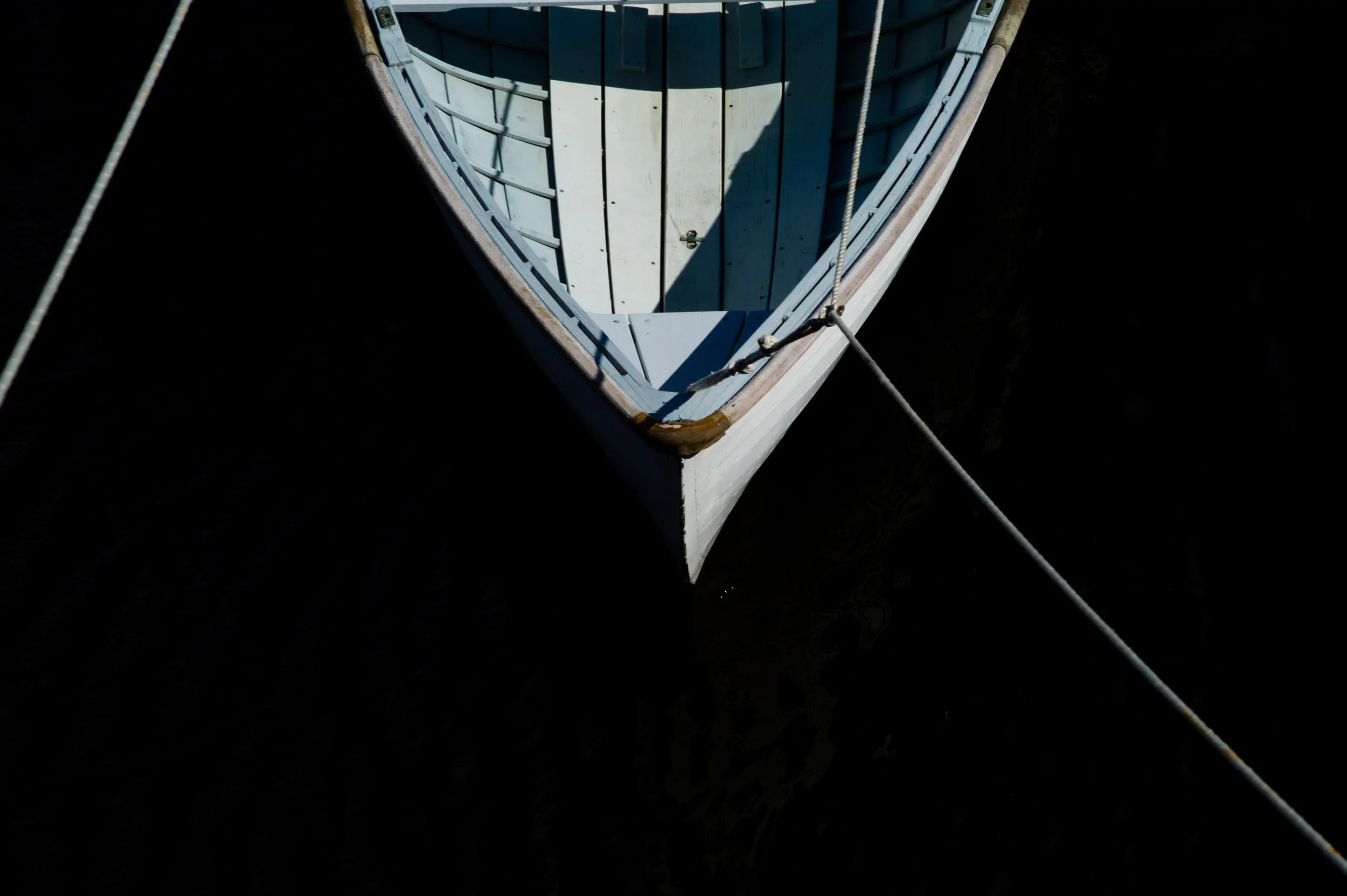 Top-down view of a white wooden boat moored in dark water, with ropes attached to the bow.