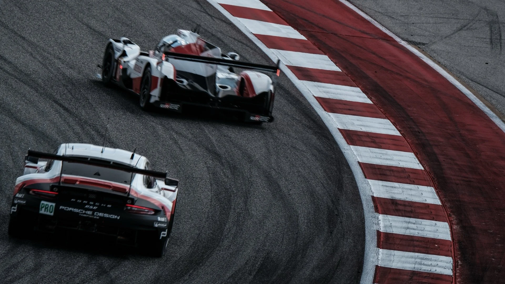 Two racing cars on a race track, taking a sharp left turn on a curve with red and white striped curbing.