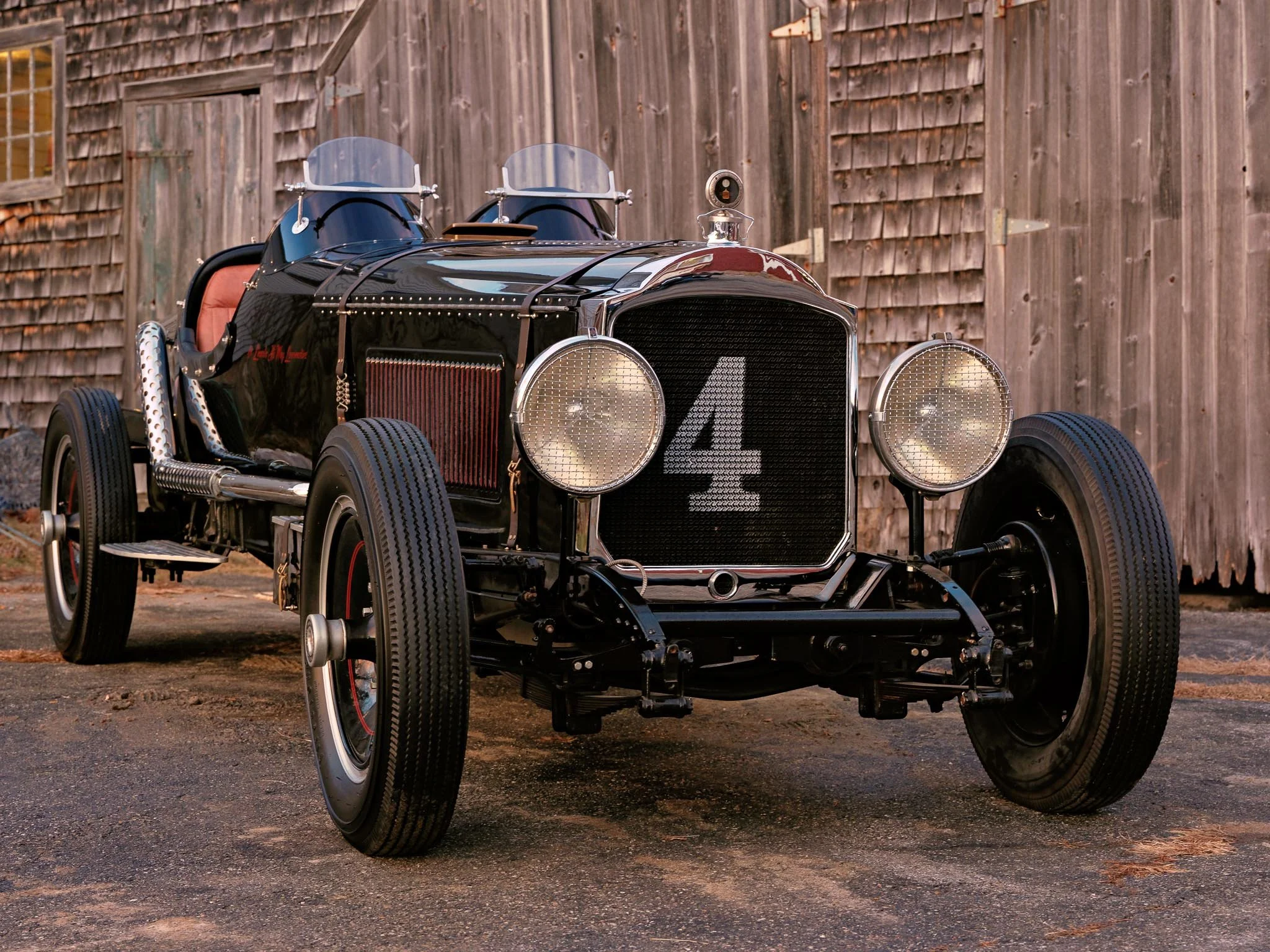 Vintage black race car with the number 4 painted on the front grille, parked on a gravel surface in front of a wooden barn.
