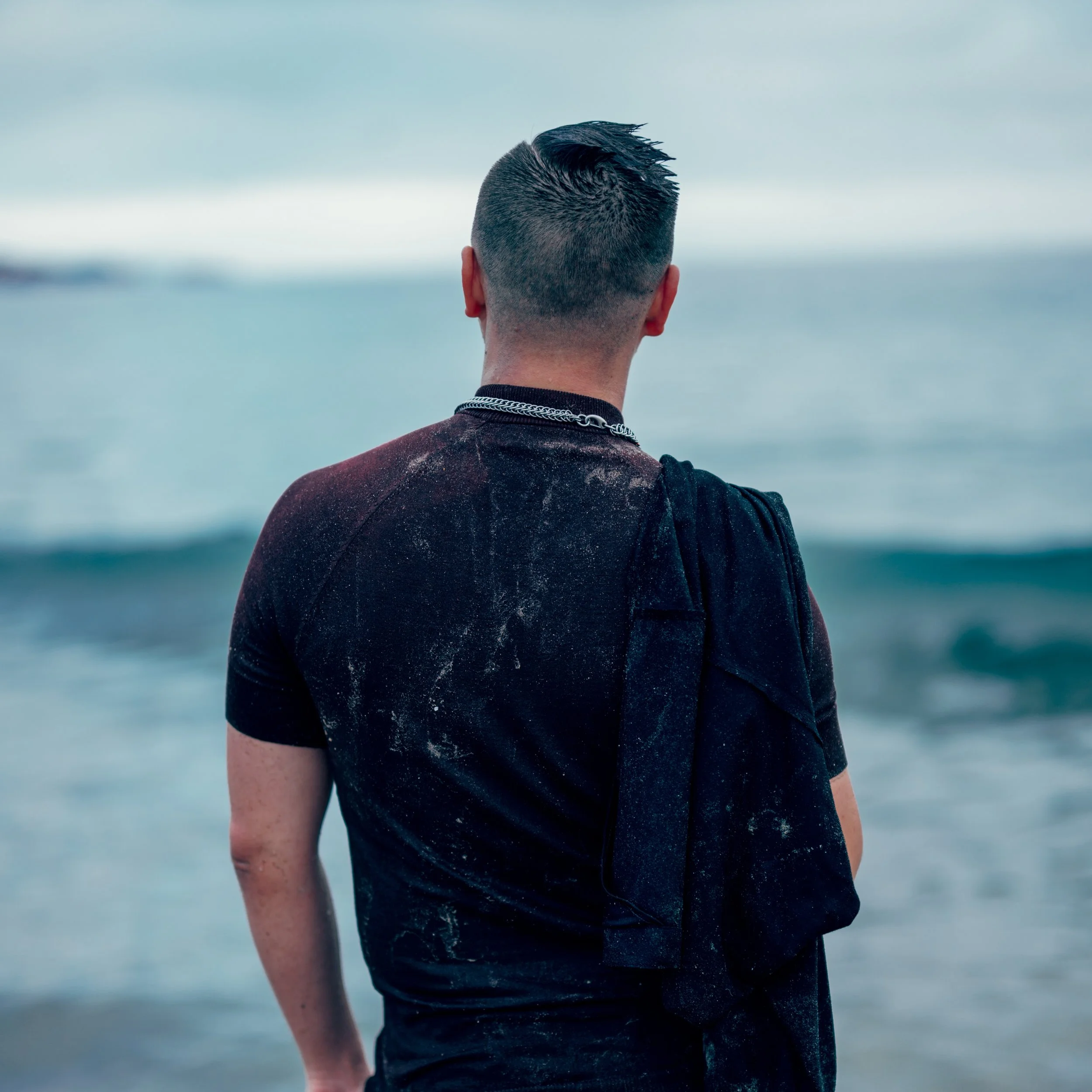 Back view of a person standing on the beach facing the ocean, with short dark hair, black shirt, and a black backpack.