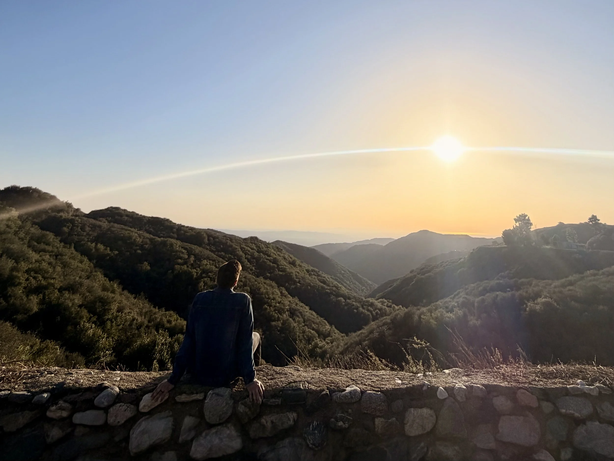 Person sitting on a stone wall overlooking a valley and mountains at sunset.