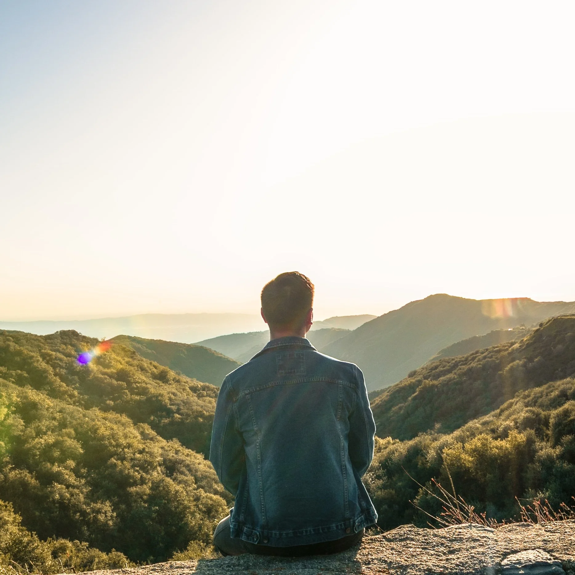 Person in a denim jacket sitting on a rock, overlooking green mountains during sunset.