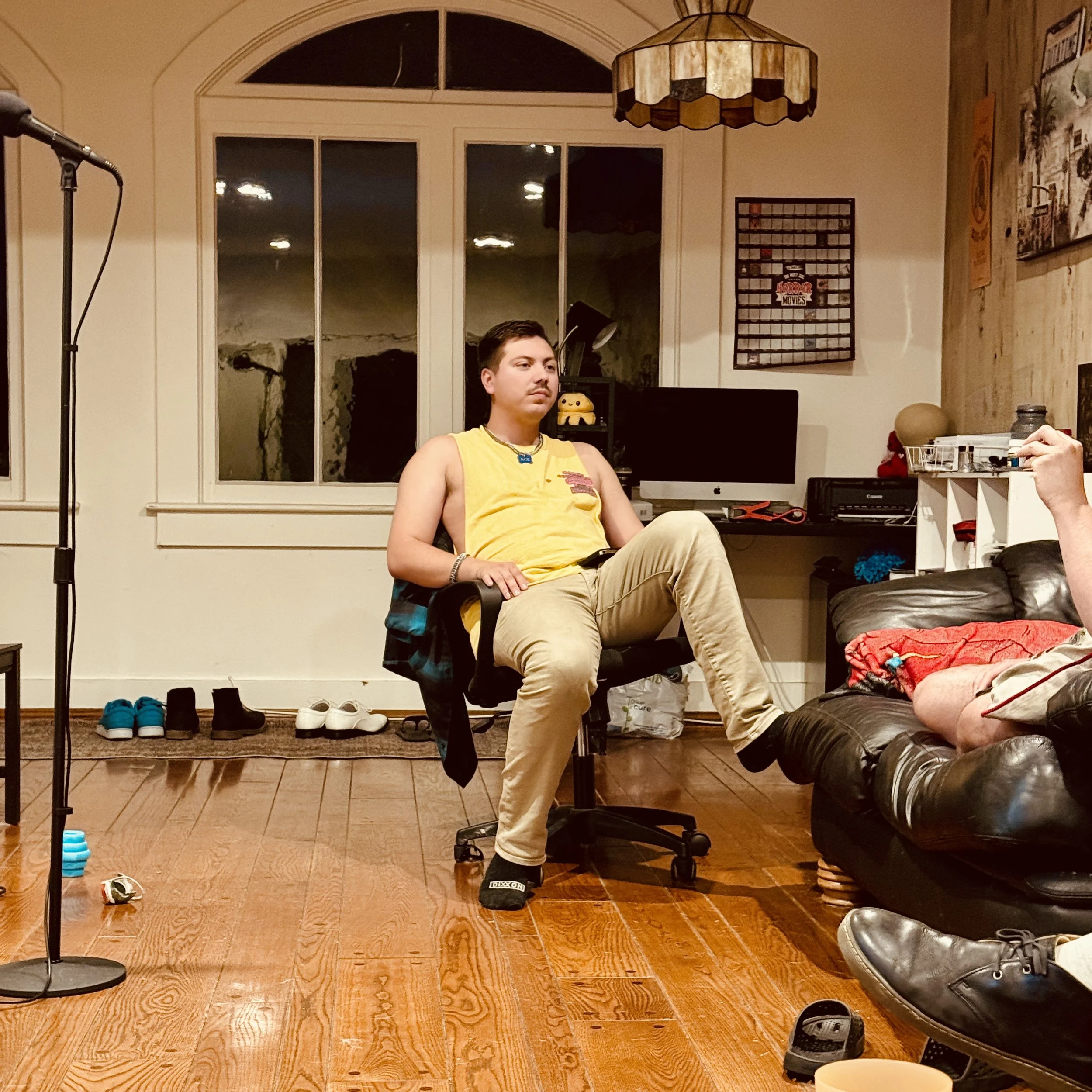 A young man sitting in a black office chair in a living room with wooden floors, wearing a yellow sleeveless shirt and beige pants, looking to his left. Behind him, a window with white trim and a view of a dark night outside. To his right, a black desk with a computer monitor and various items. On the floor, there are shoes and a small toy or object. To his left, a black microphone stand is visible. Part of another person is seen on the right side, sitting on a black leather couch, with part of their arm and a red blanket visible.