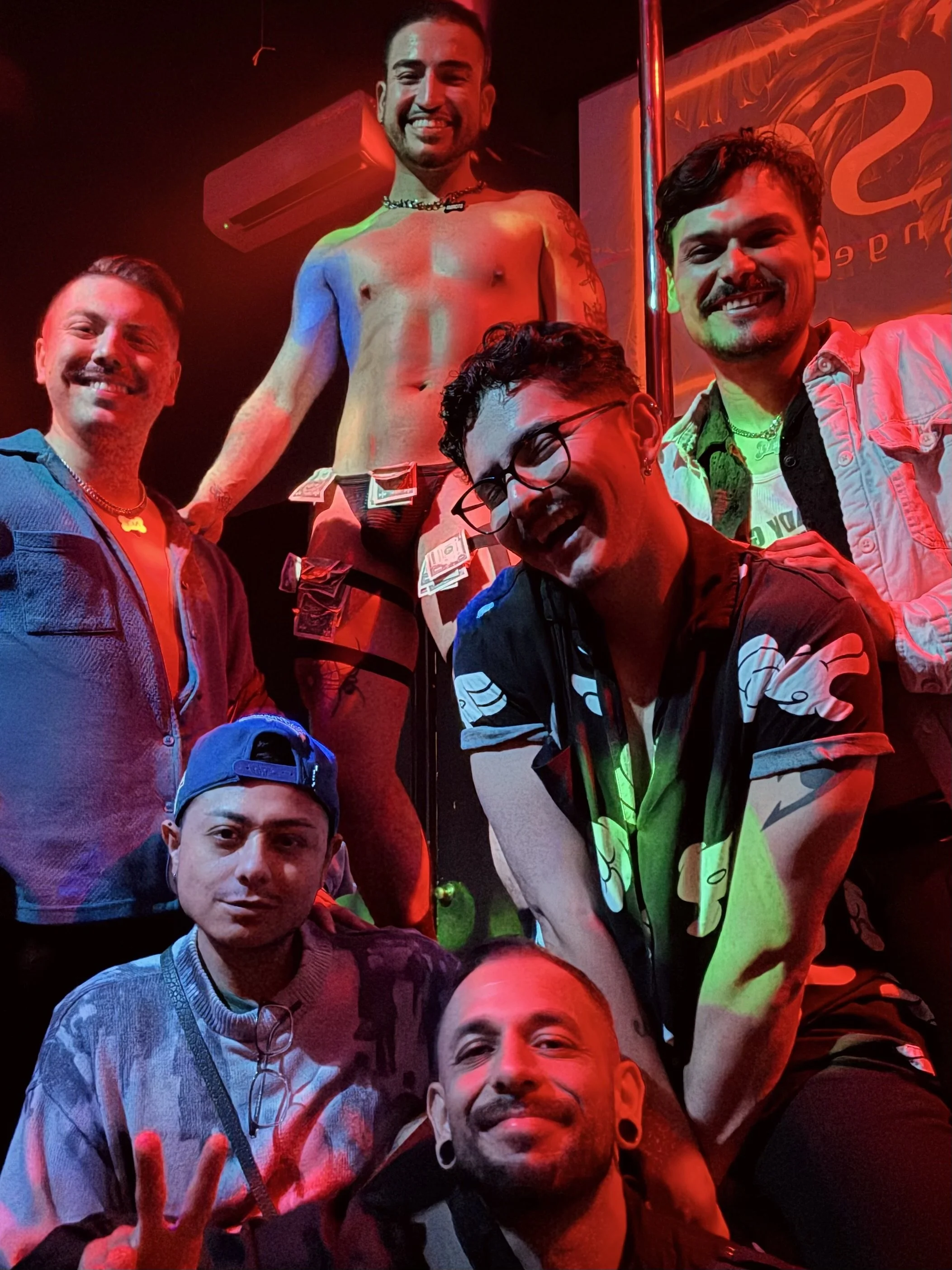Group of six friends posing together in a bar with colorful lighting, one person shirtless with dollar bills on his waist, and a neon sign in the background.
