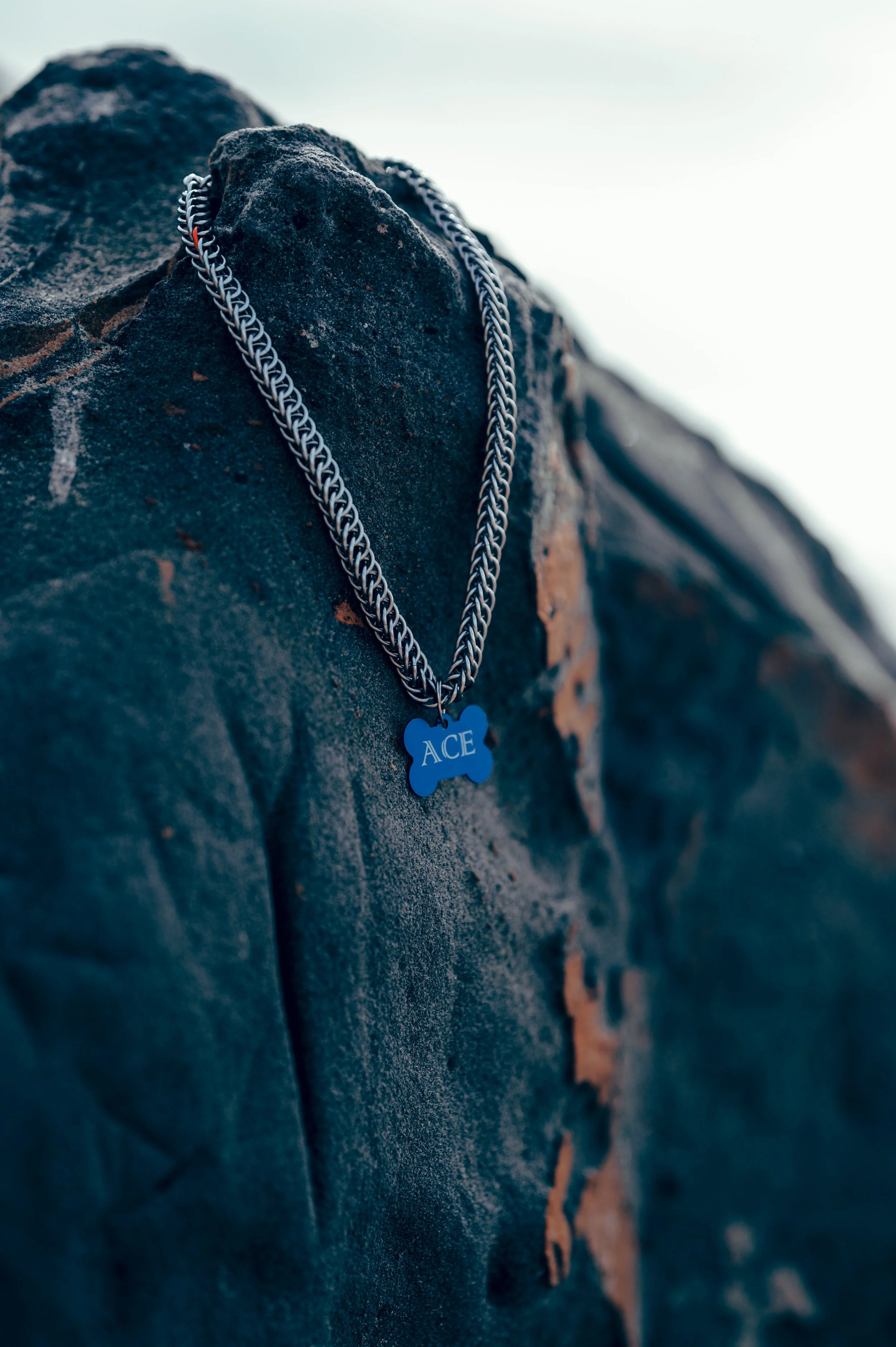 A silver chain necklace with a blue bone-shaped pendant engraved with the name 'ACE' hangs on a textured black rock.