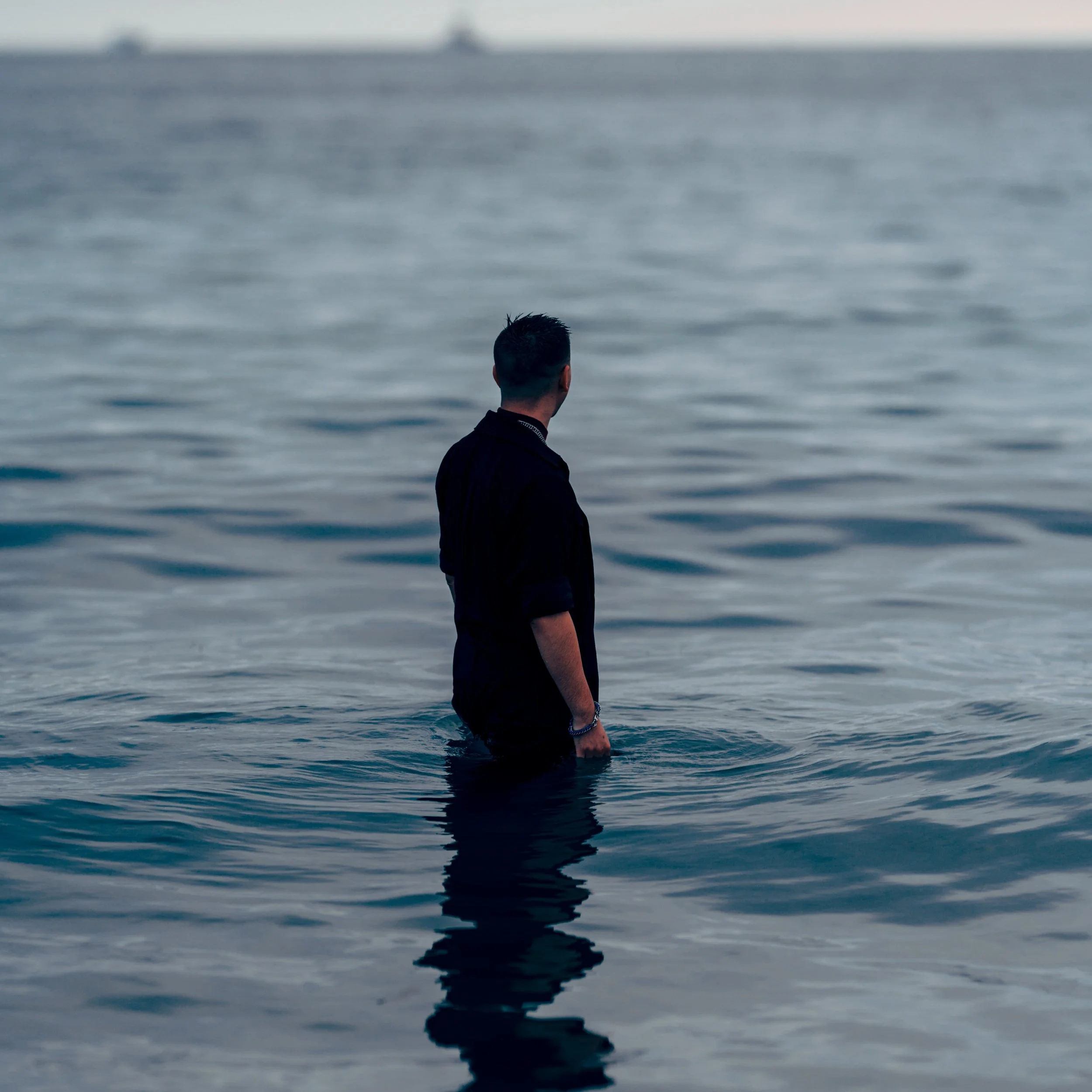A man standing in the ocean, seen from behind, wearing a dark shirt with water up to his waist.