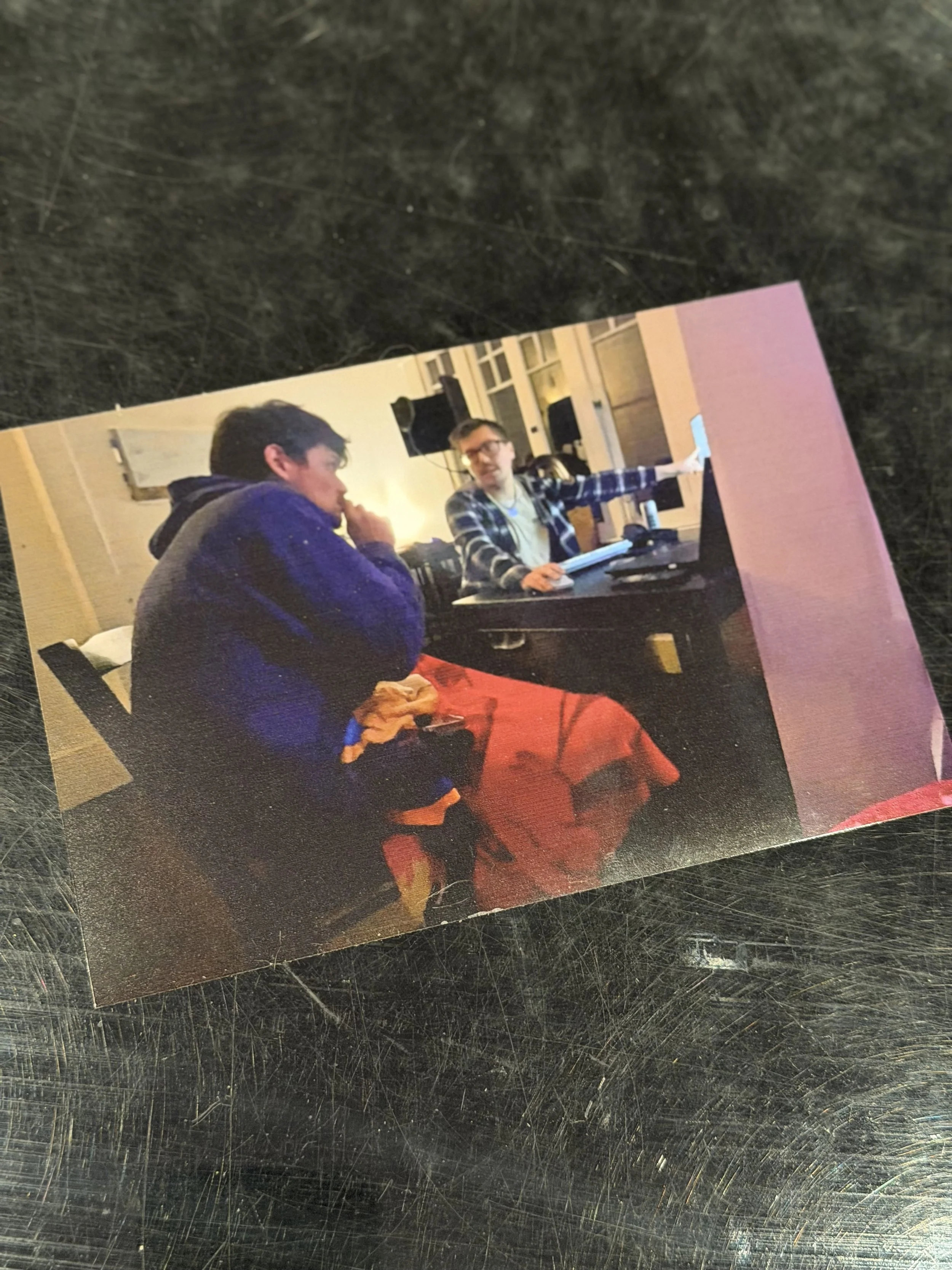 Two young men are sitting at a desk, engaged in a discussion. One is wearing a blue jacket and the other a checkered shirt, with a computer and equipment in a room with large windows.