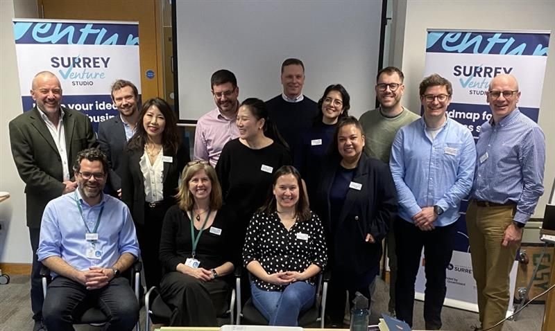 Group of 14 people standing and sitting in a conference room, smiling for a group photo during a workshop or seminar. Two banners with 'Surrey Venture Studio' are in the background.