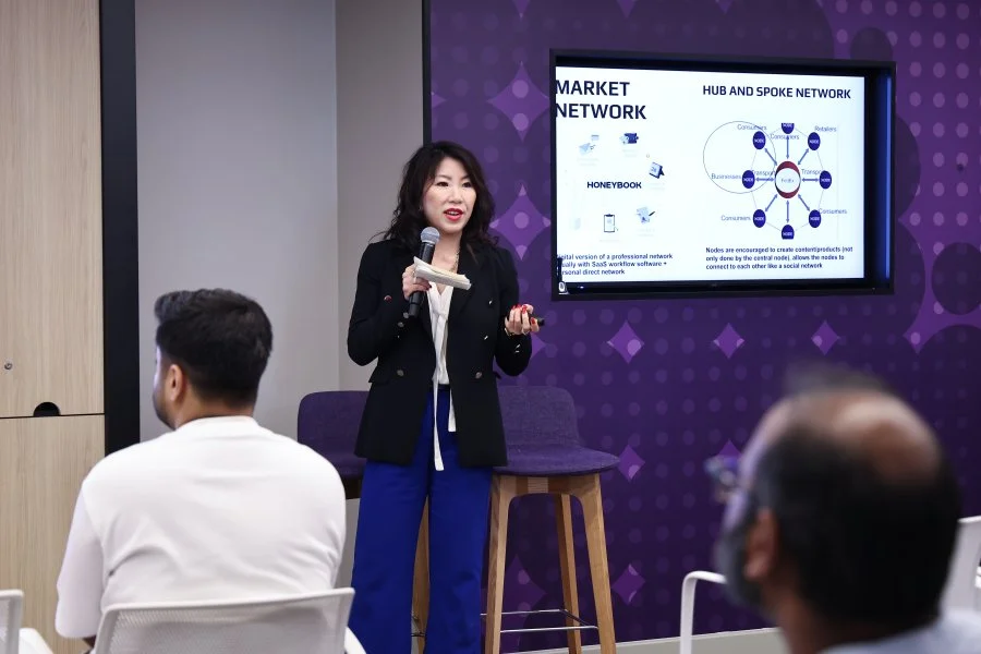 A woman giving a presentation in a conference room with a purple patterned wall, holding a microphone and a remote. A screen behind her displays a slide on market network, hub and spoke network, and a diagram of interconnected nodes. Attendees are seated and listening, one person with glasses and another with dark hair and a white shirt.