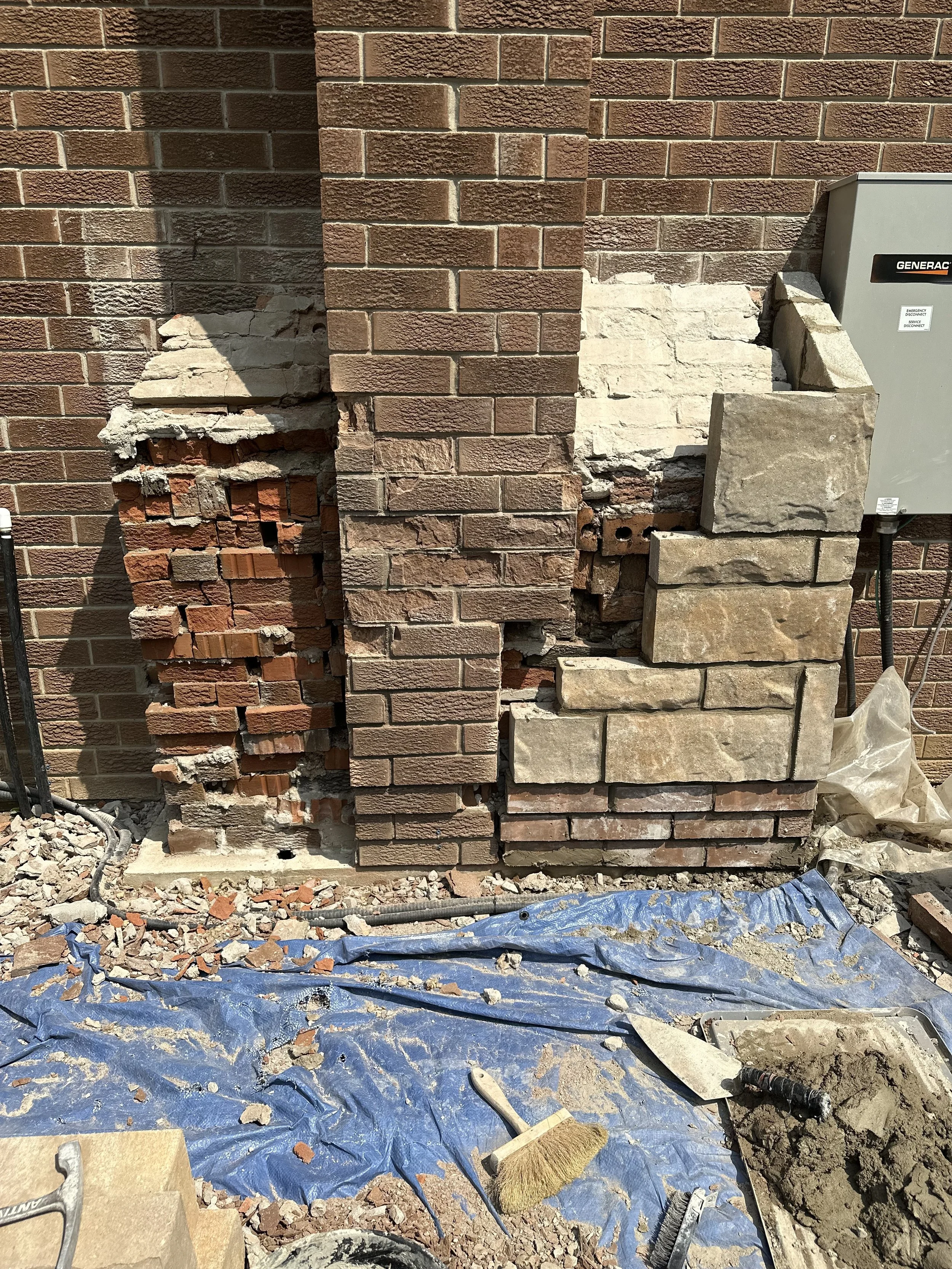Construction site with damaged brick wall, tools, and debris on blue tarp.