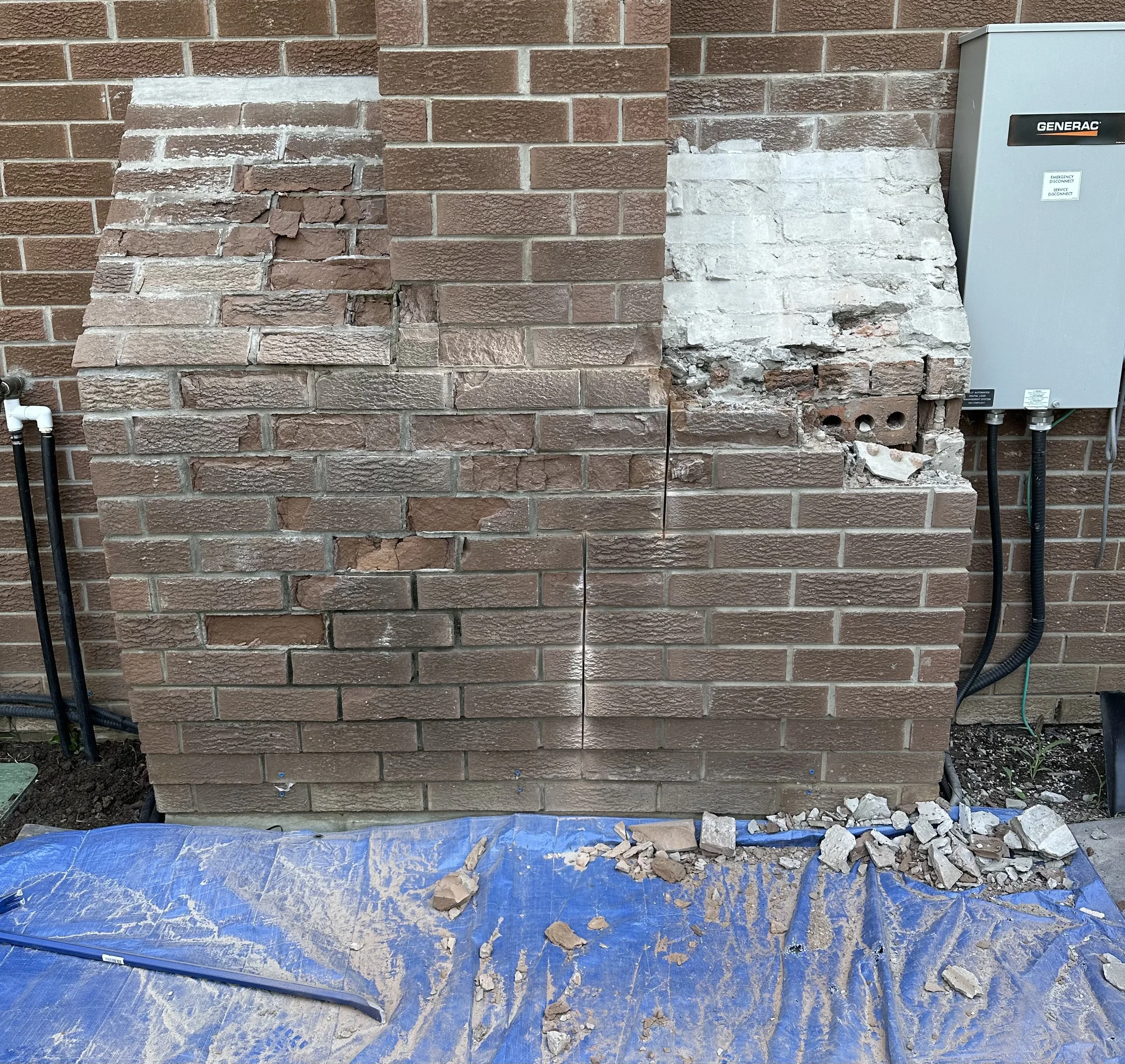 Section of brick wall with part of damaged bricks removed, exposing the underlying structure. Construction debris and broken bricks on a blue tarp on the ground.