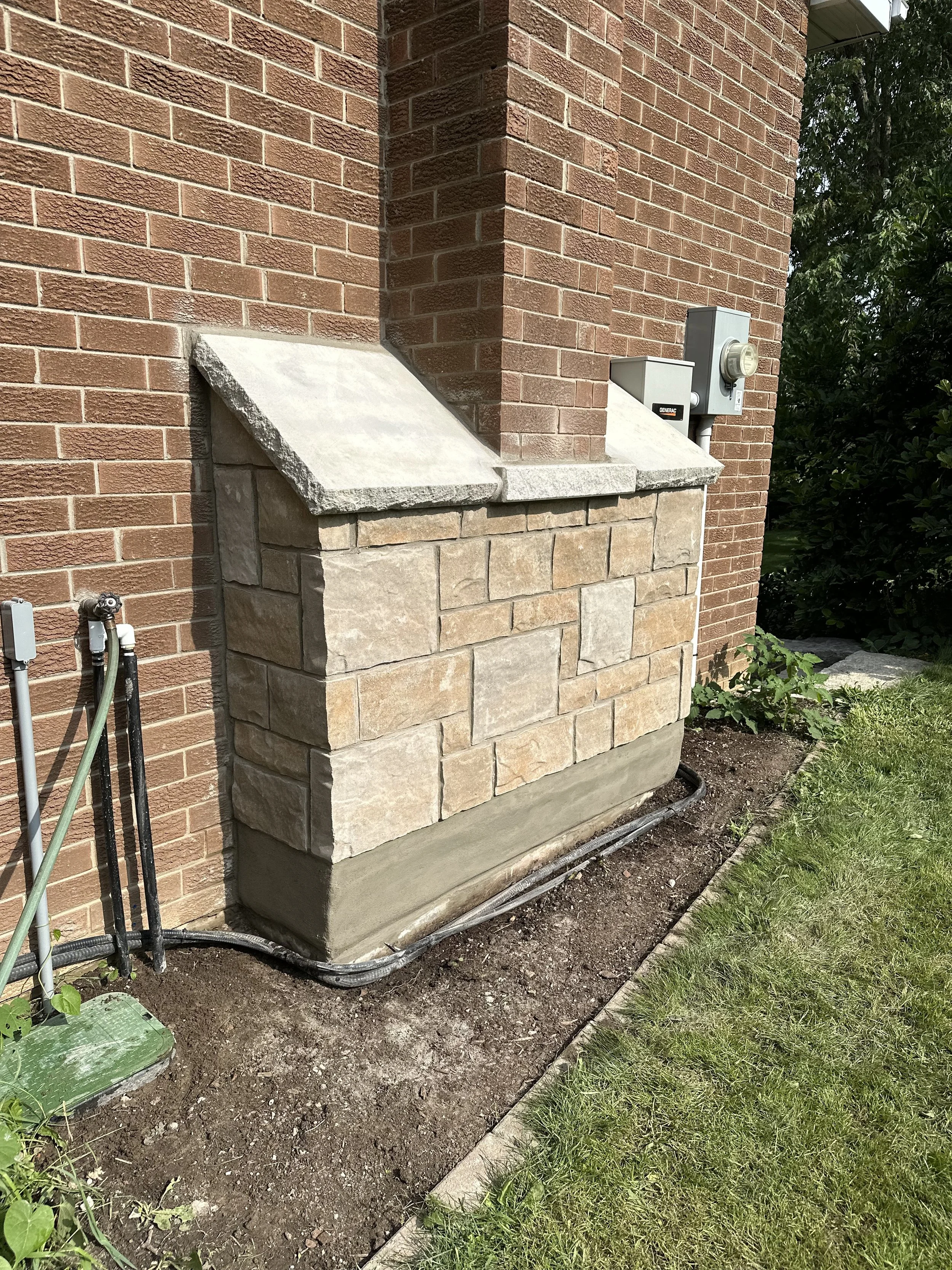Close-up of a brick exterior wall with a stone planter and electrical meters or boxes mounted on it, and a garden hose outlet nearby.