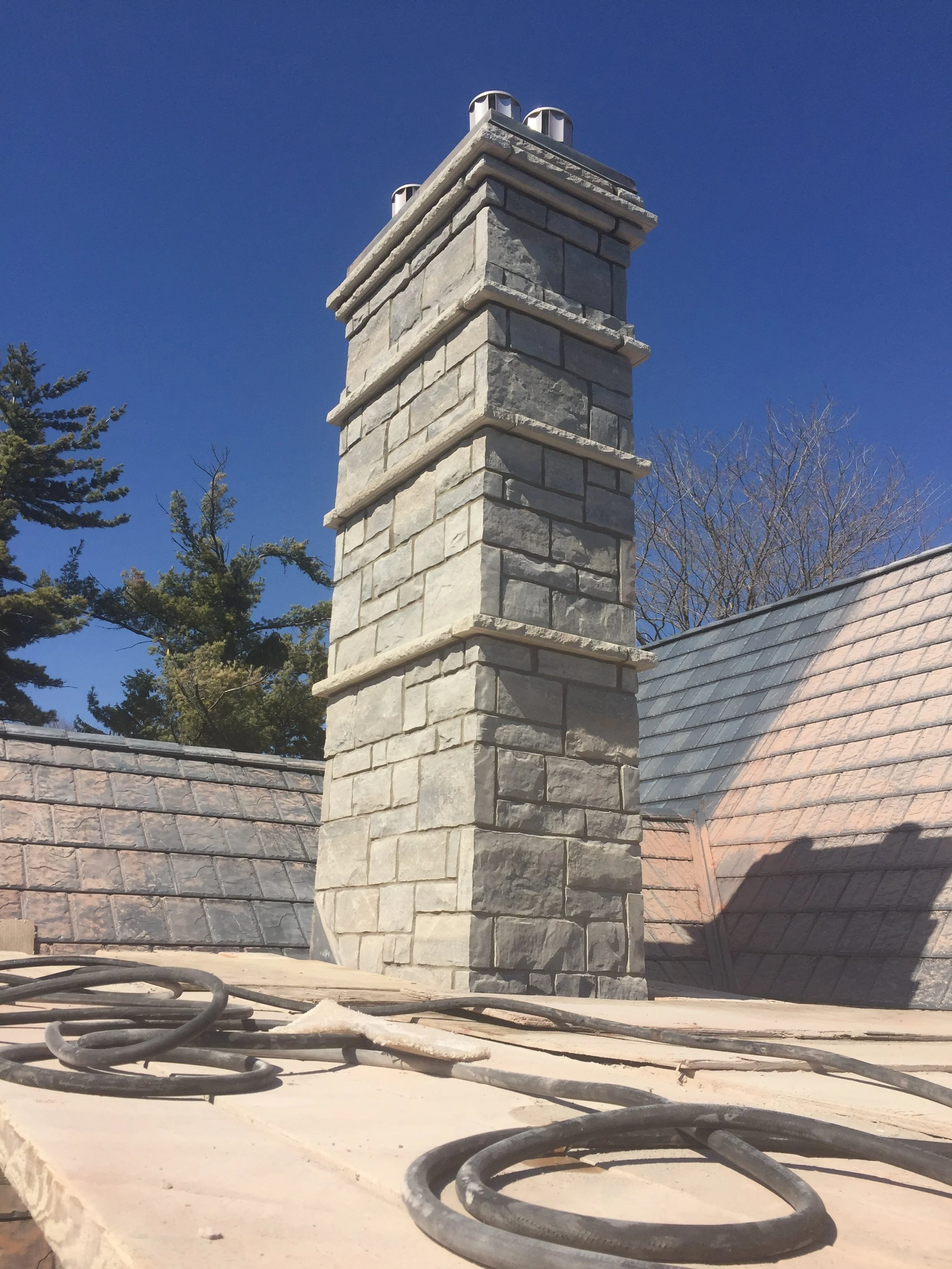 Close-up view of a chimney made of stone bricks on a rooftop under a clear blue sky, with some black cables and equipment in the foreground.