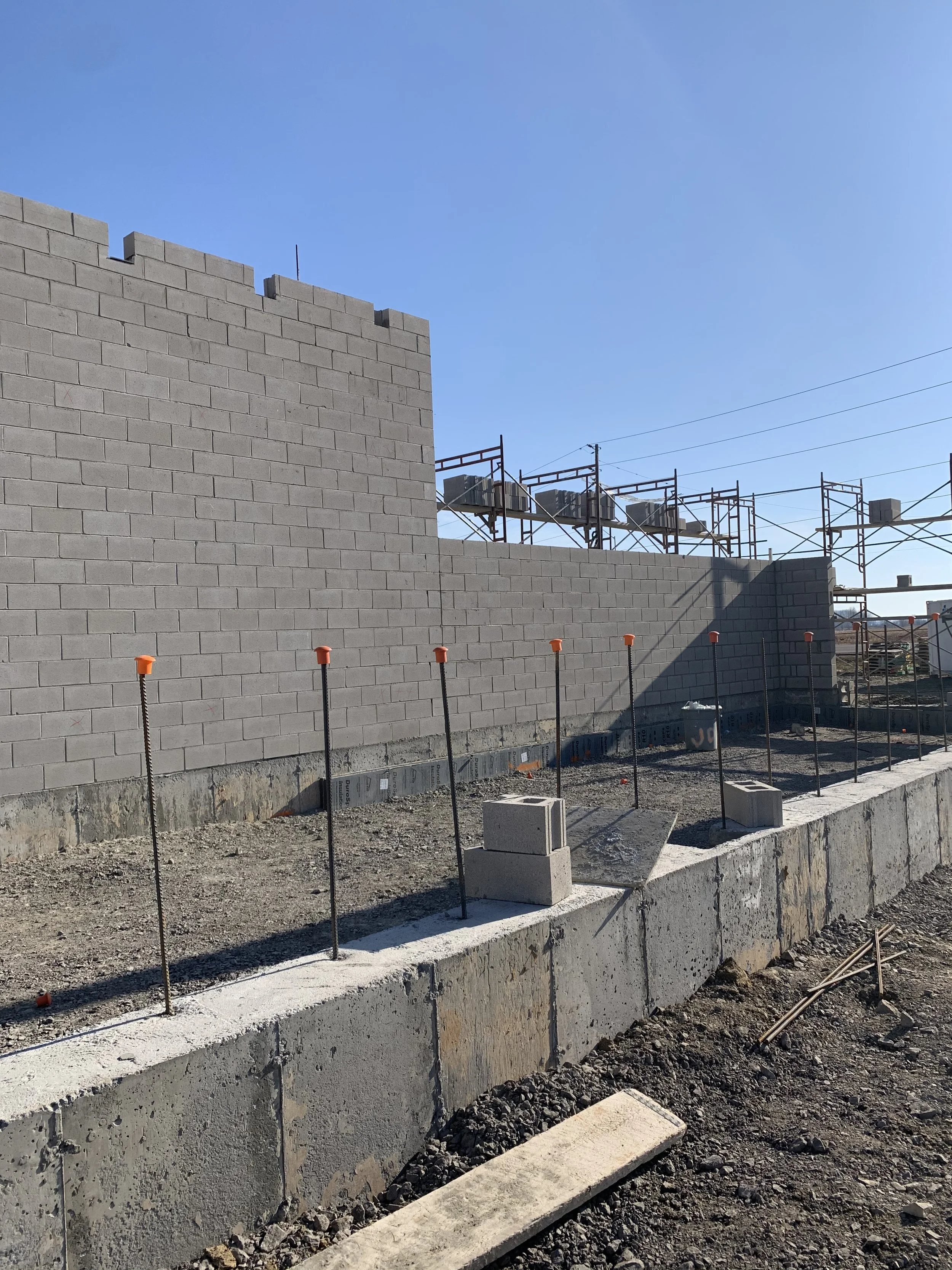 Construction site with gray cinder block walls, orange safety markers, and scaffolding under a clear blue sky.