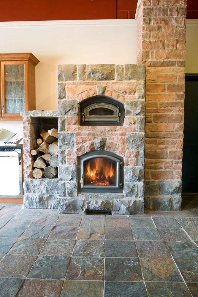 Stone fireplace with a wood-burning stove at the bottom, a small arched door for firewood on the side, and a metal oven door above the stove in a cozy living room.