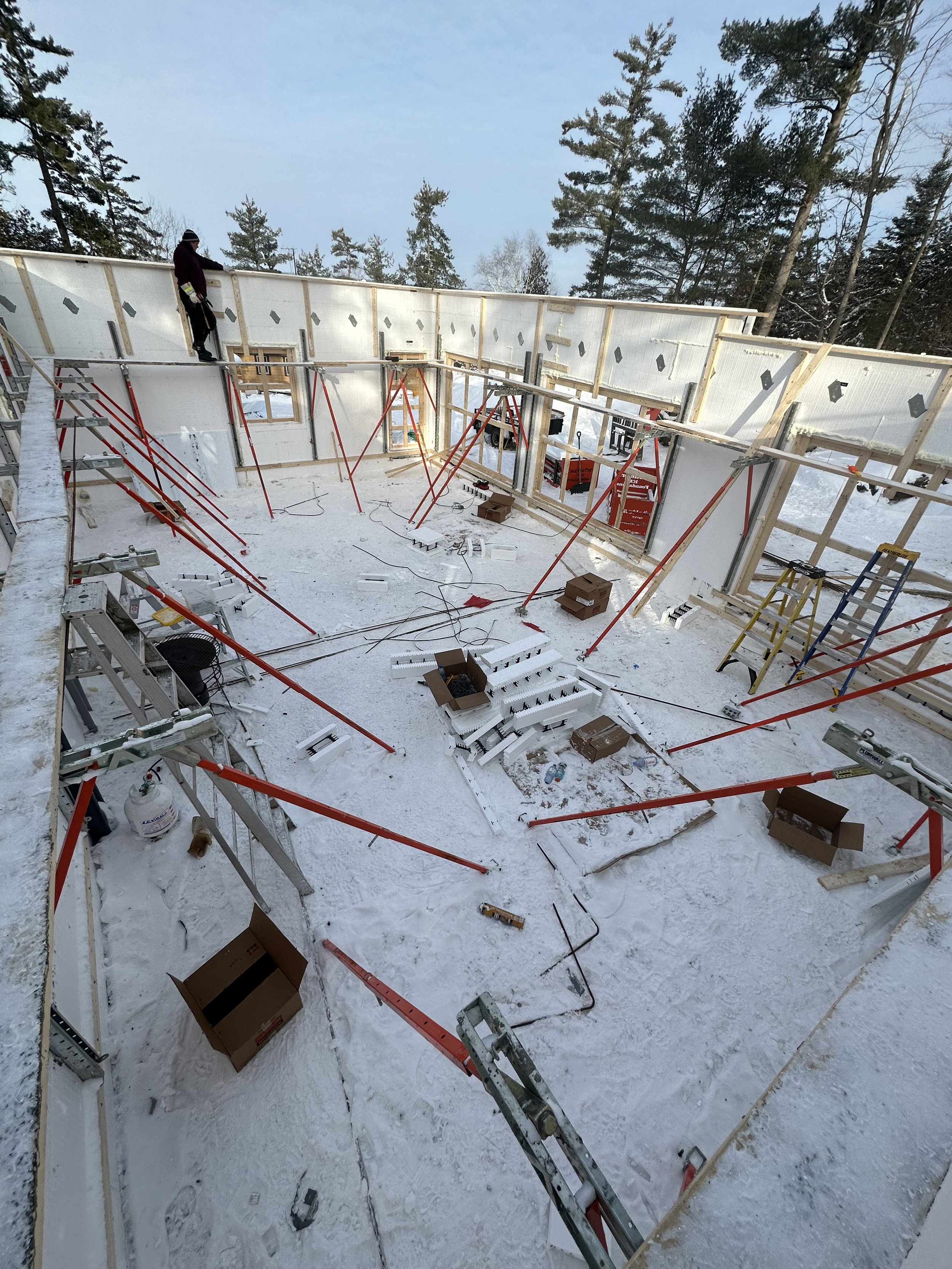 Construction site in winter showing the framing of a house with a person standing on the top of the wall, surrounded by snow, ladders, and building materials.