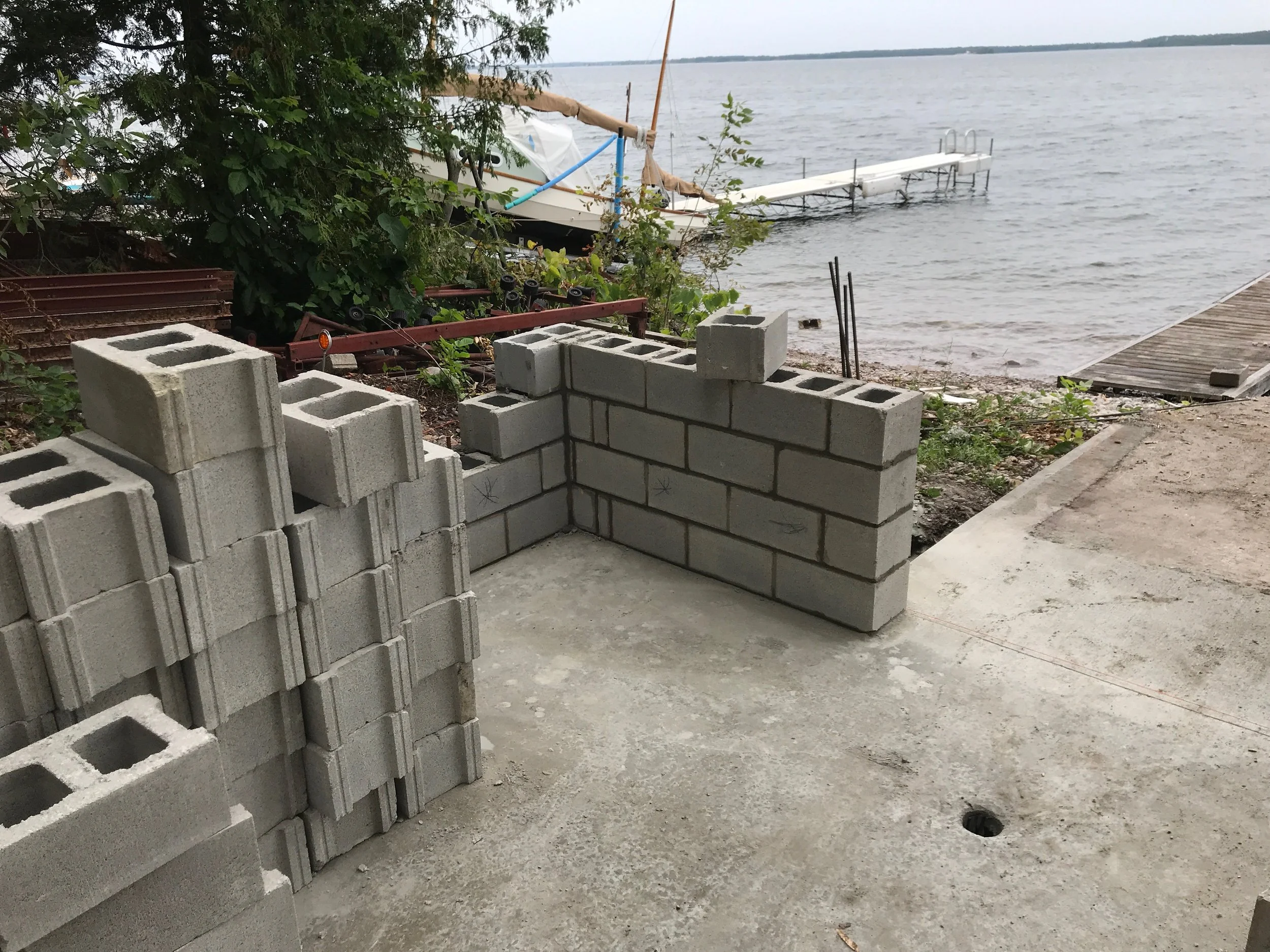 Construction site near a body of water with stacked concrete blocks and a partially built wall, a dock extending into the water, and trees in the background.