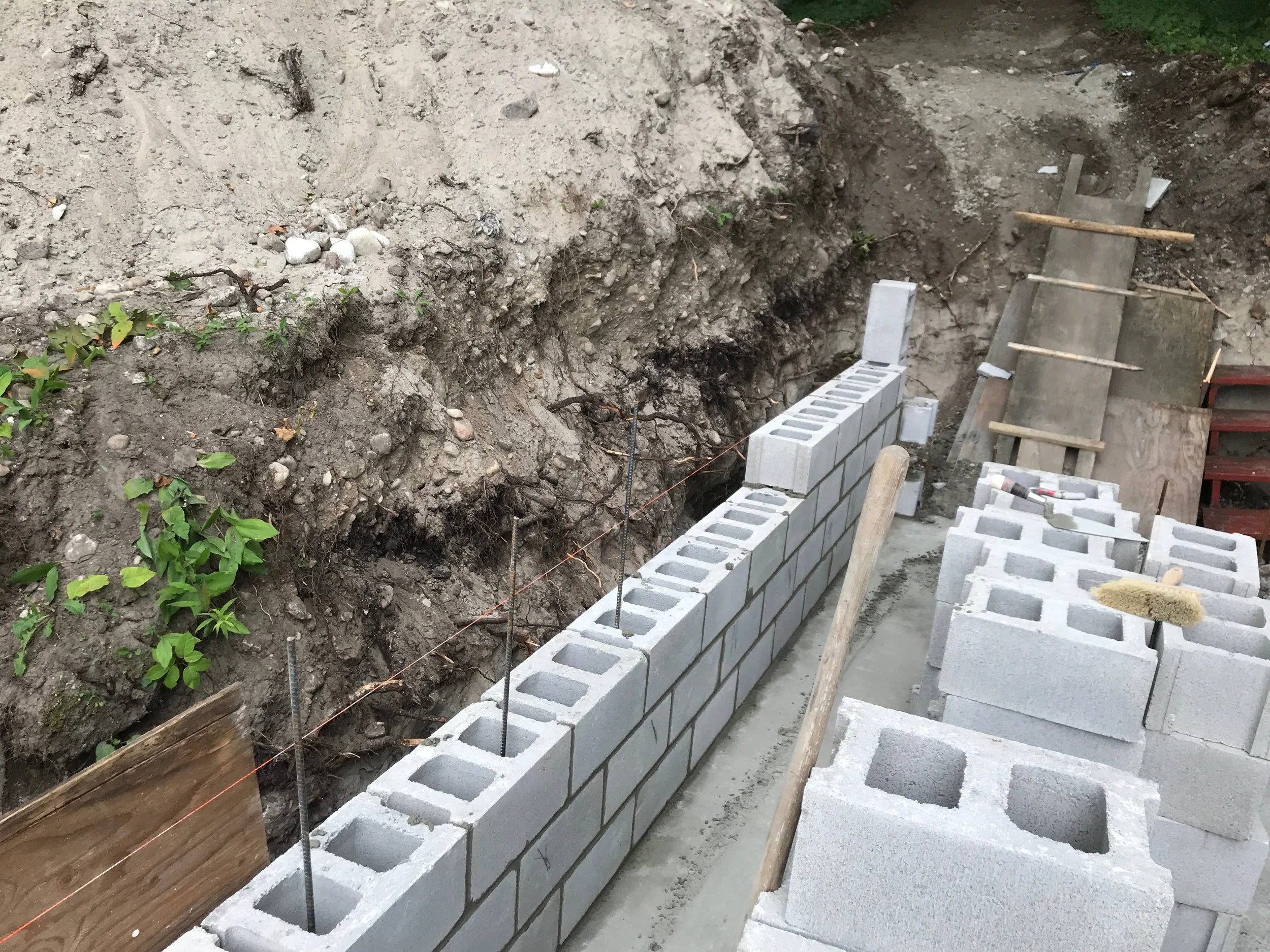 Construction site with a row of concrete cinder blocks being laid for a wall, with tools and wooden planks nearby, and a dirt trench behind it.