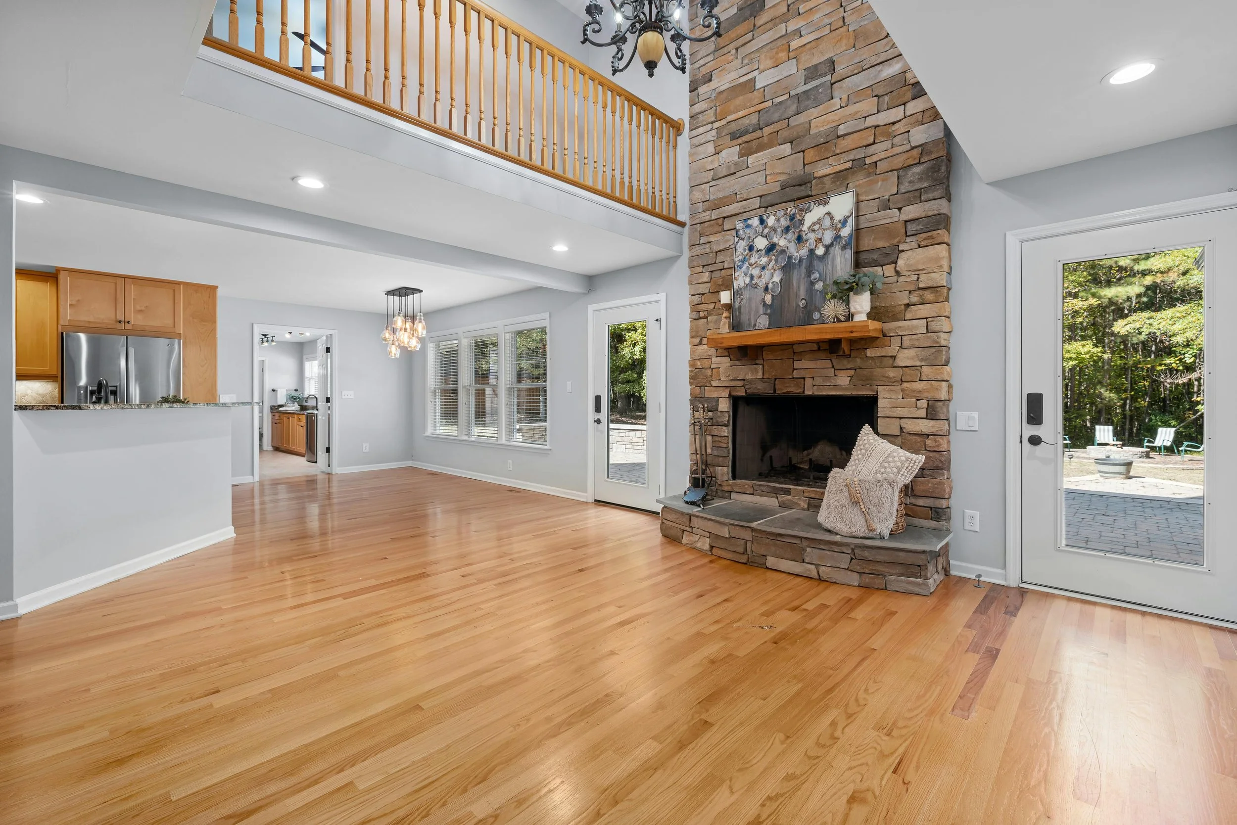 Living room with a brick fireplace, a wooden mantel decorated with plants, and a couch. Large windows and glass door lead outside to a patio with outdoor furniture. Hardwood floors and a chandelier hanging from the ceiling.