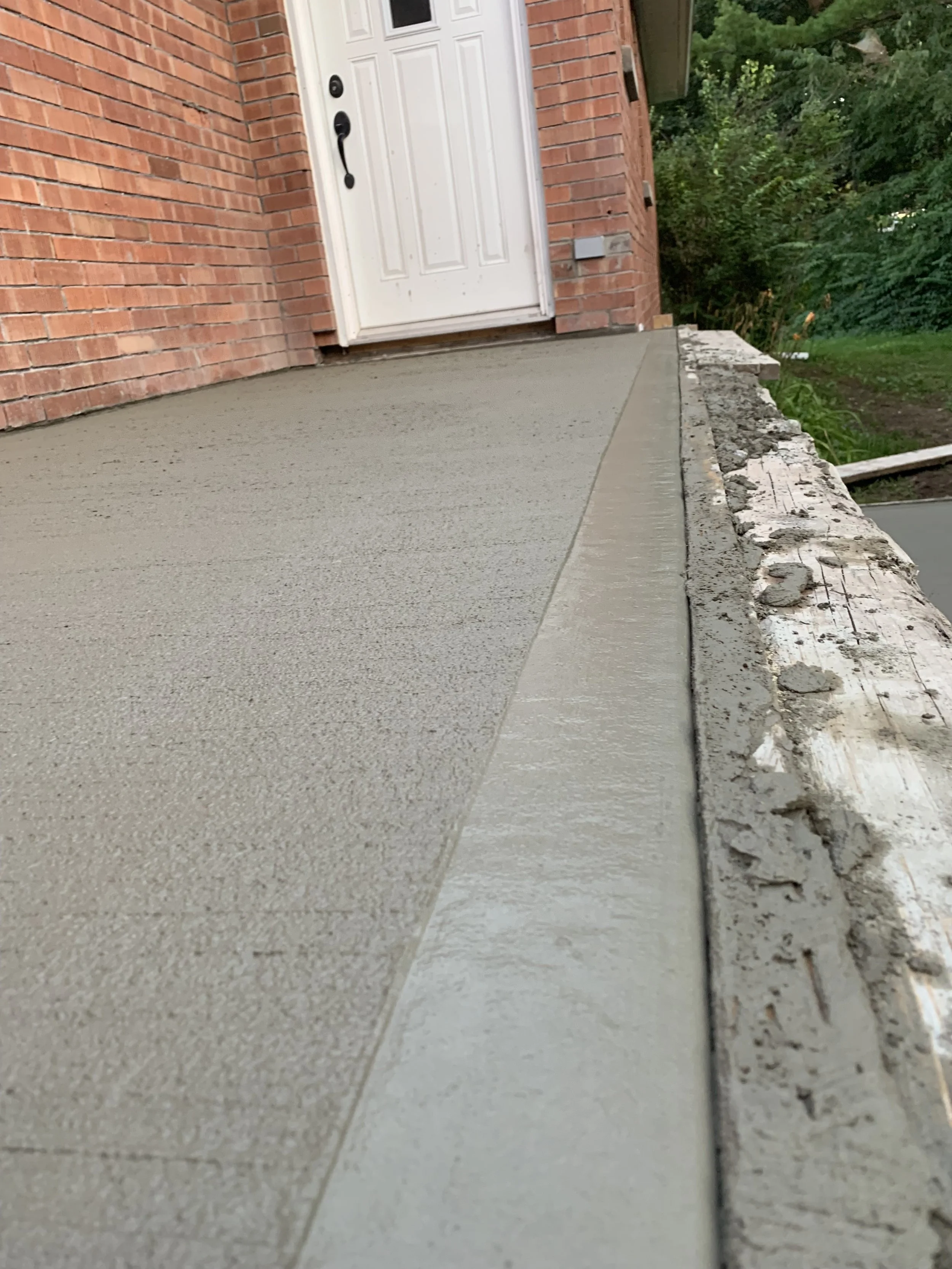 Front porch with new concrete surface and brick wall of house, white door, and surrounding greenery.