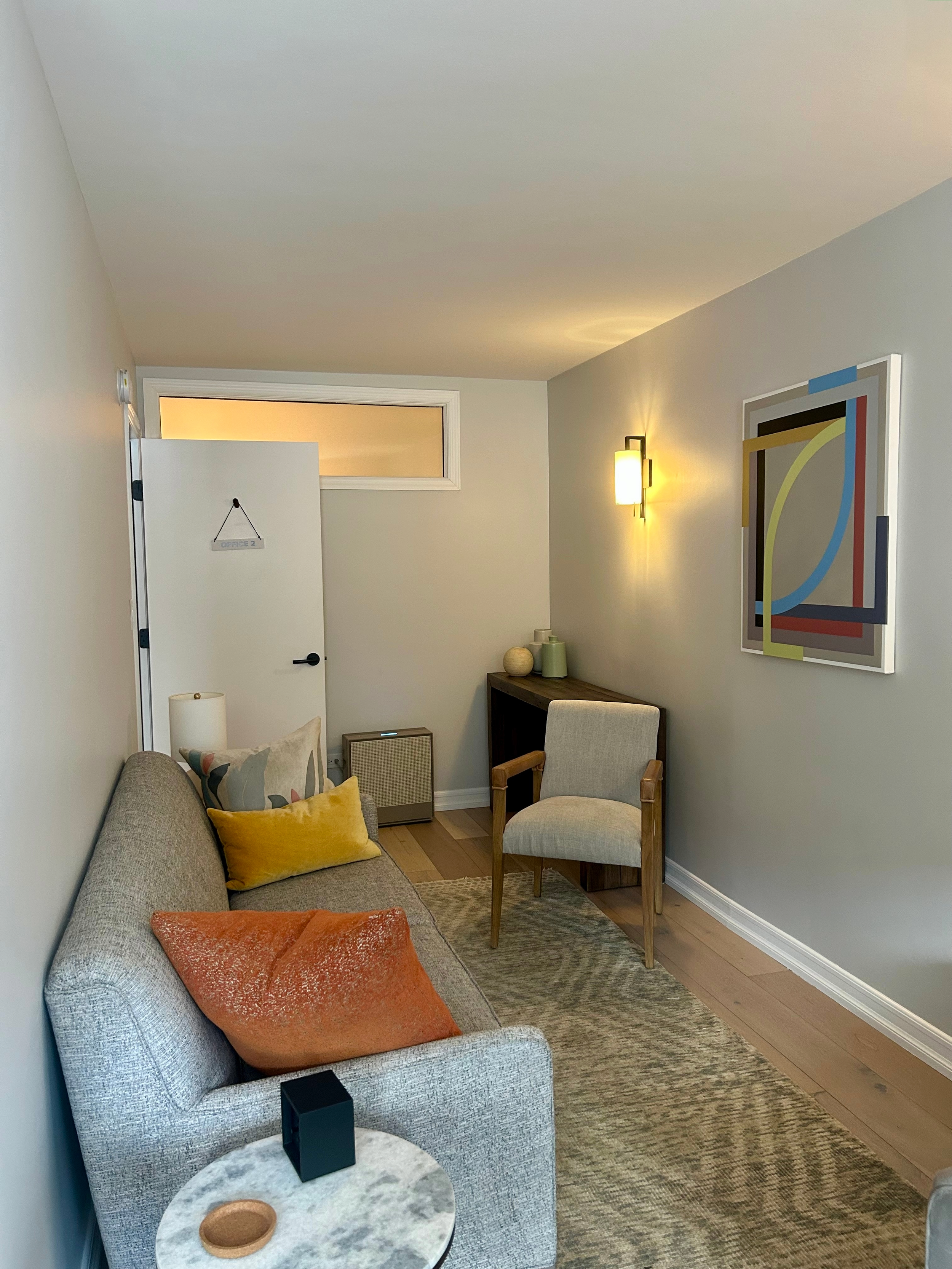 Office Two in a therapy office for rent in Brooklyn, with a sofa, pillows, a small round marble coffee table, a beige armchair, wall art, wall sconce lighting, and a wooden side table with decorative vases.