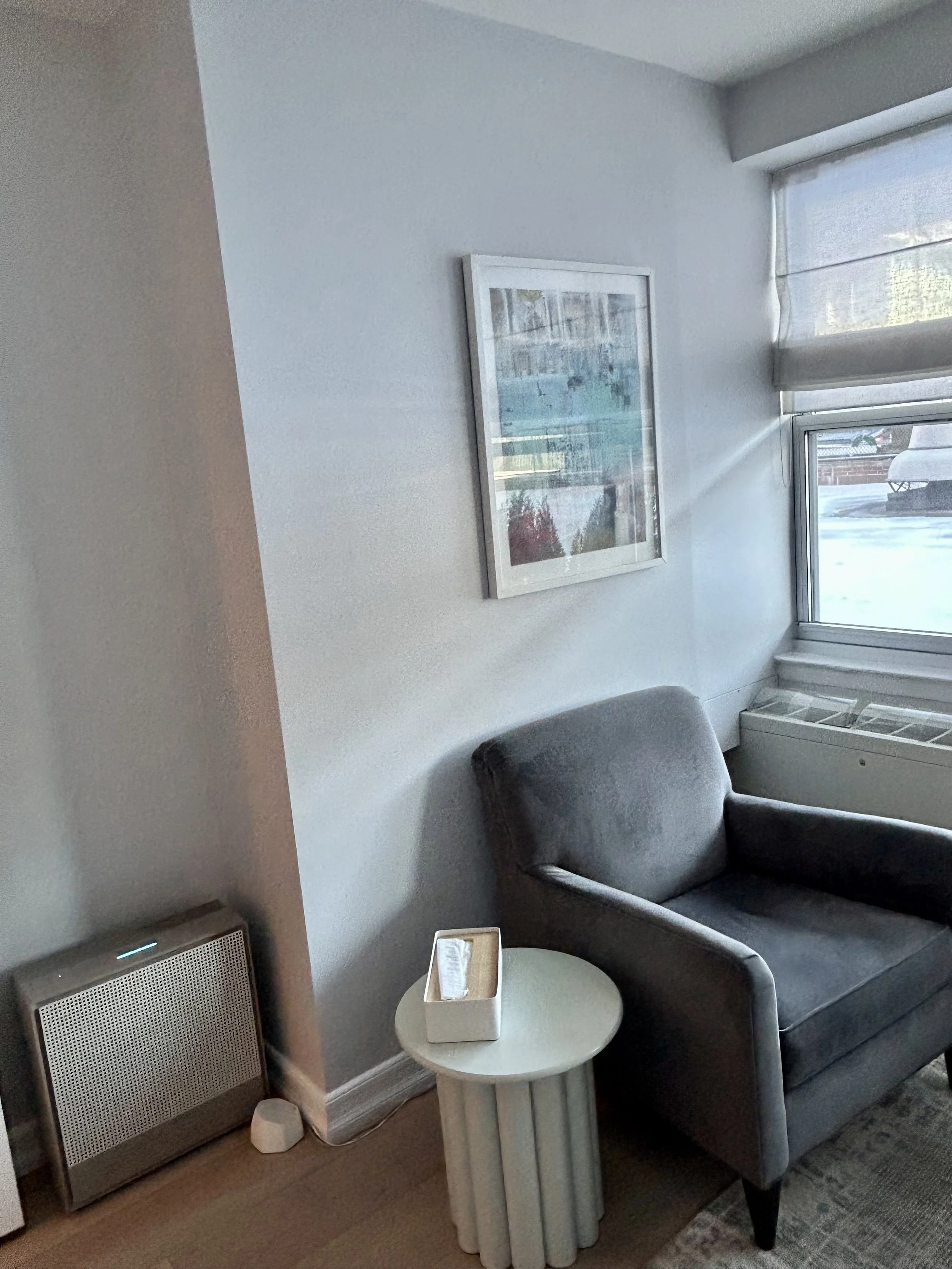 A  corner of Office One, a therapy office for rent in Brooklyn, with a gray armchair, a white round side table with a tissue box, a painting on the wall, a window with a Roman shade, and a radiator/AC unit.
