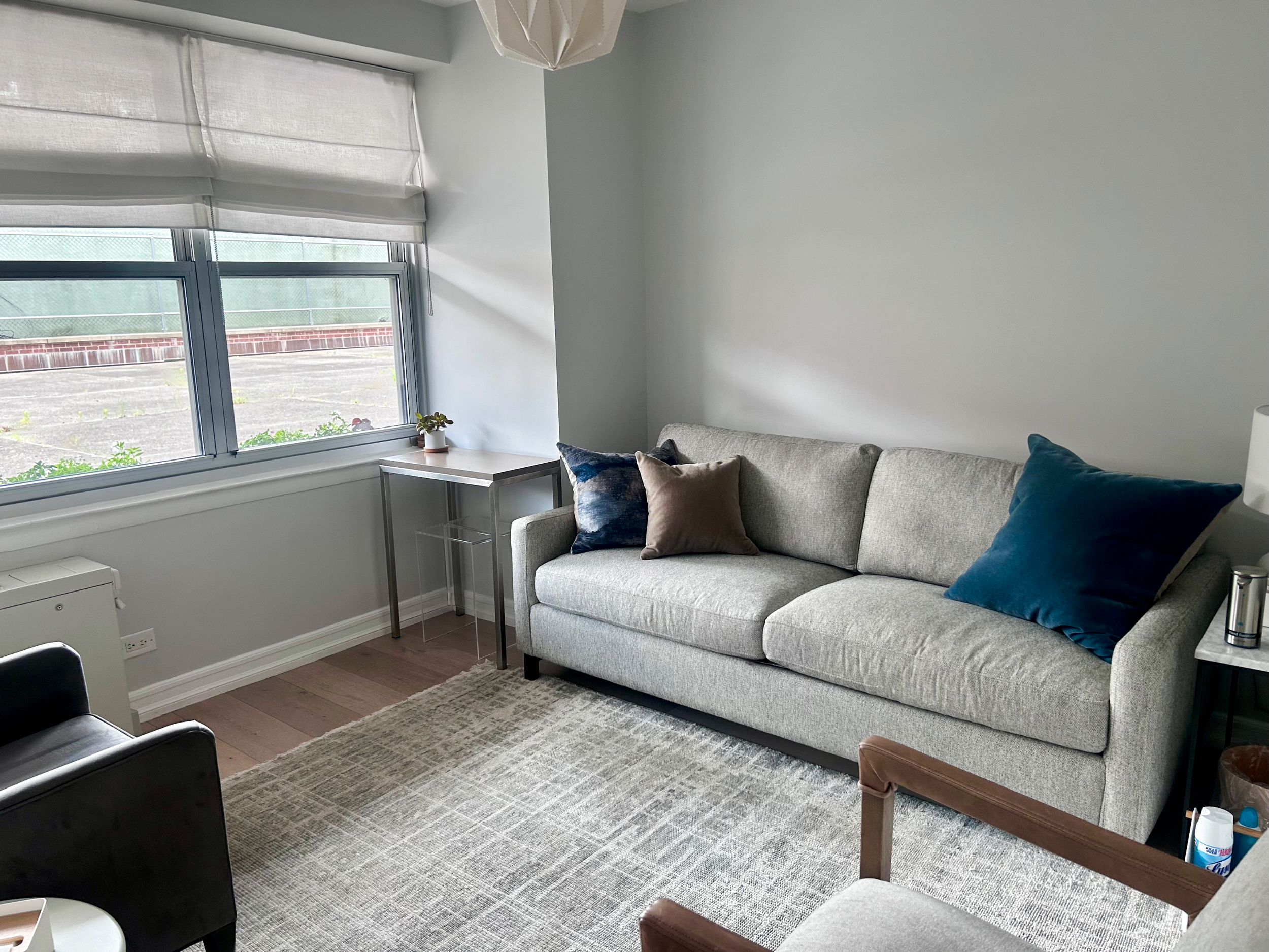 Office One in a therapy office for rent in Brooklyn, with beige sofa, navy and beige pillows, small side table, window with Roman Shade, potted plant,  area rug, and part of an armchair.