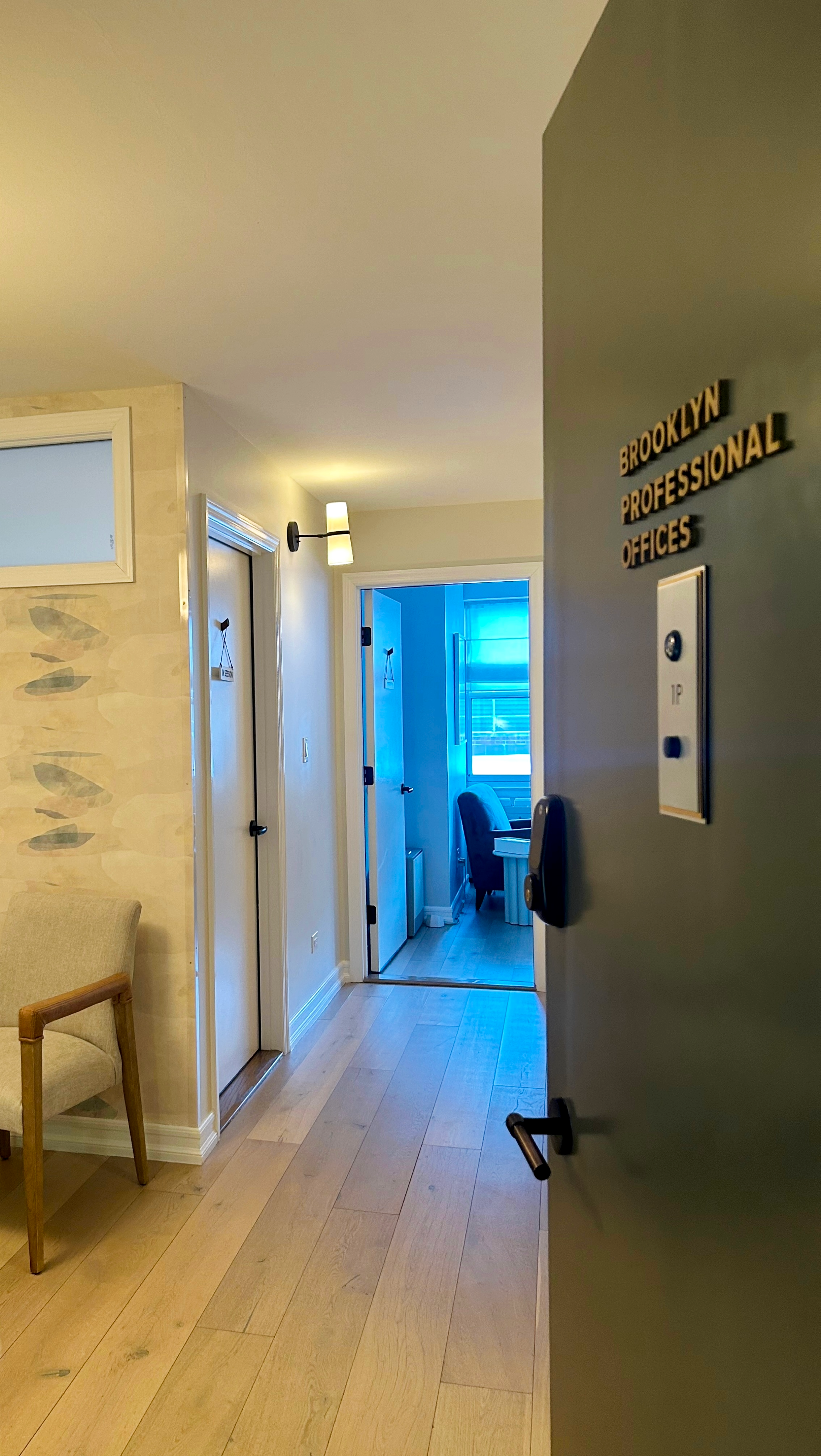 View of Brooklyn Professional Offices' entry door,  leading into a  room with a window and a chair inside.  Hallway has wooden flooring, wall-mounted lights, and a sign on the door. This is the entry to therapy offices for rent in Brooklyn.