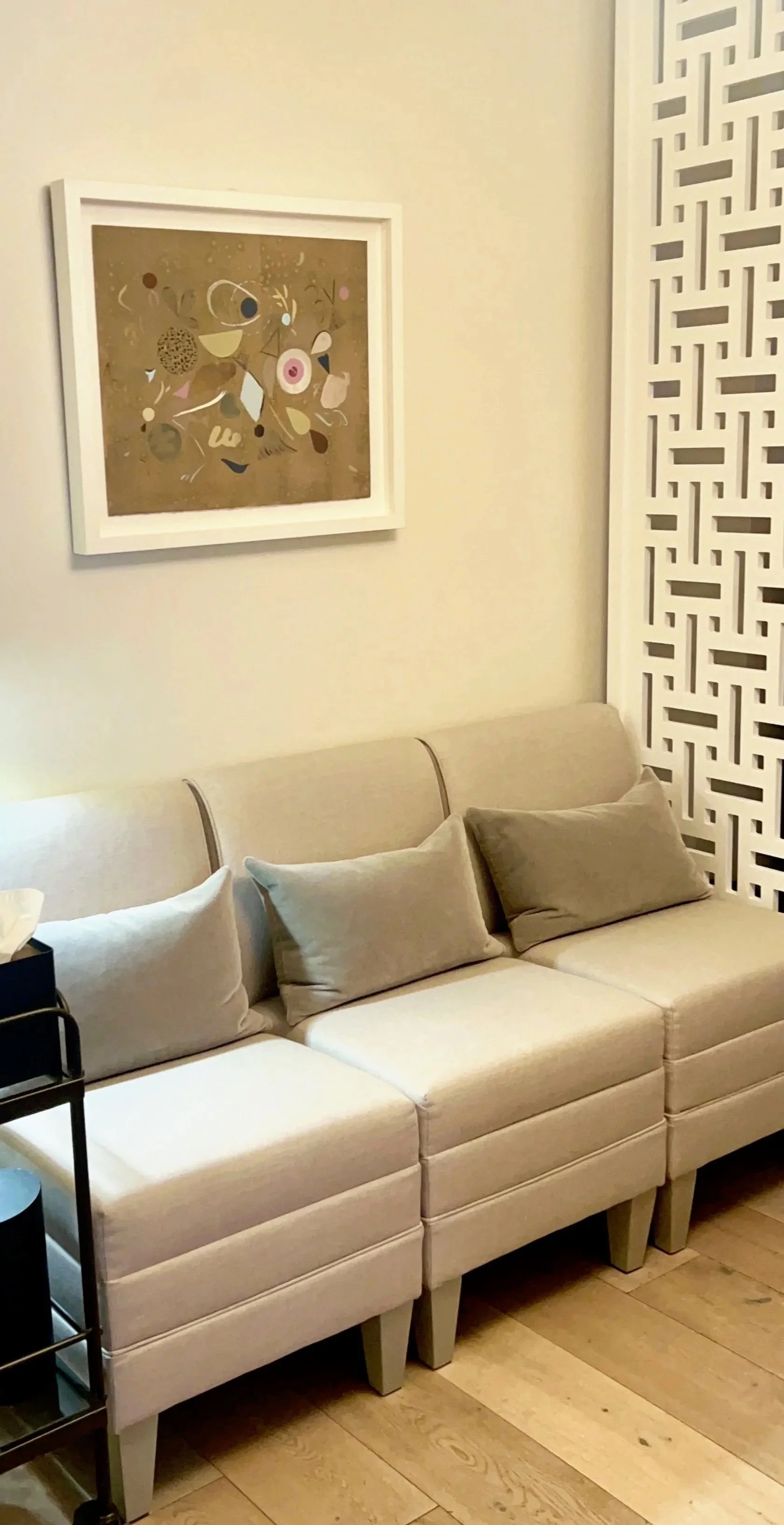 A beige sofa with three cushions in front of a white wall, decorated with a framed abstract art piece and a decorative white lattice panel.  This is the waiting room of the therapy offices for rent in Brooklyn.