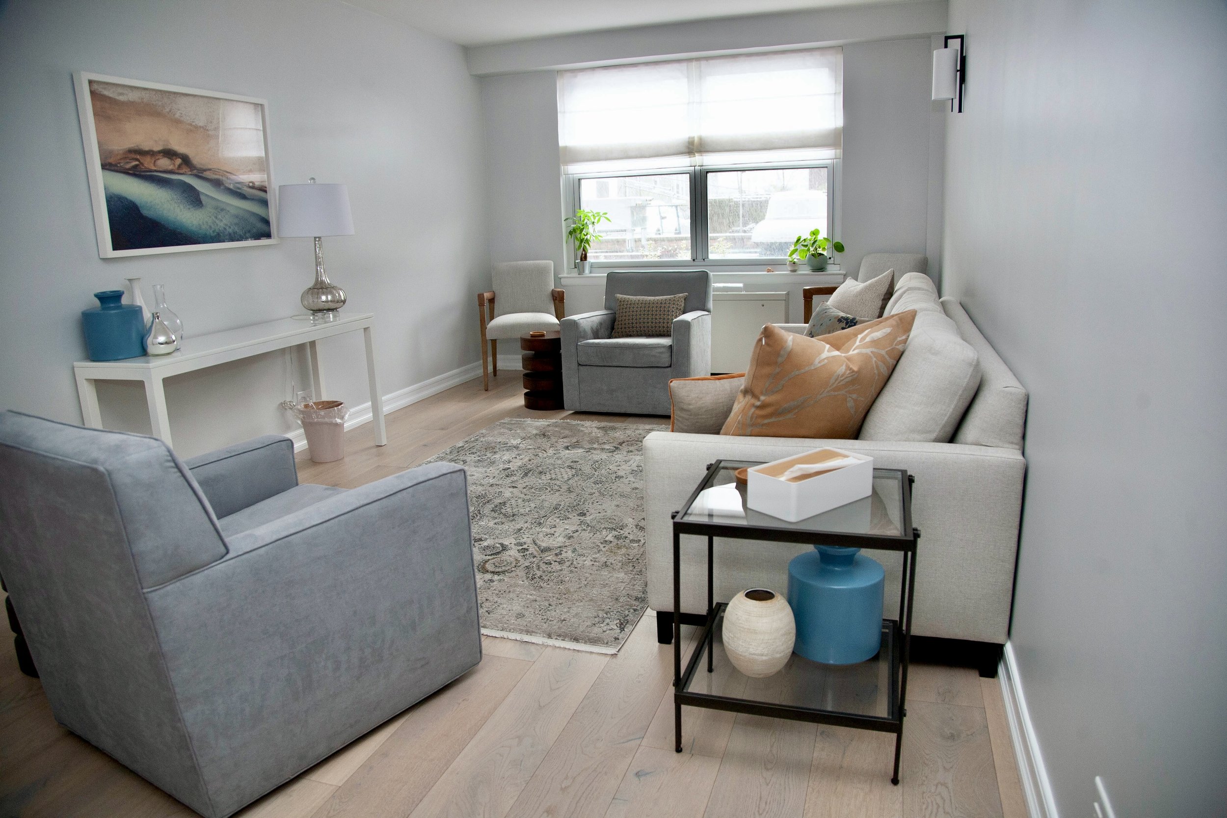 A view of Office Three in a therapy office for rent in Brooklyn, showing windows,  with gray and beige furniture, a white side table with decorative vases, and a black and glass side table with a white tissue box and vases.
