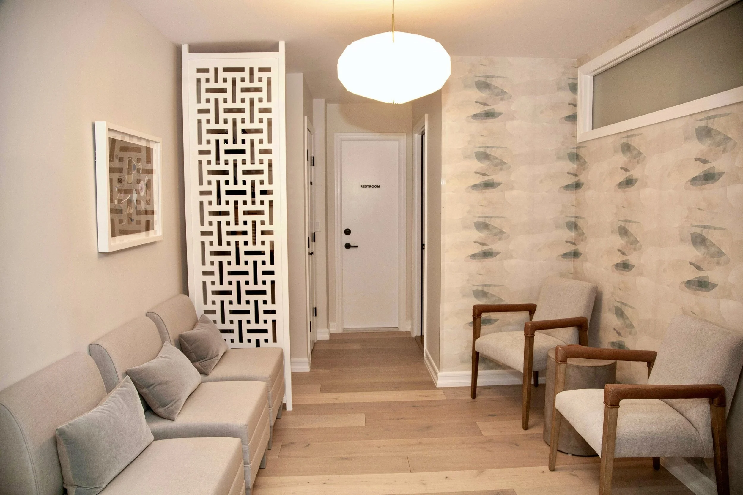 A waiting room with  beige seats with pillows, wooden chairs with upholstered seats, a decorative white partition, a framed artwork, a white door marked 'Restroom,'' This is the waiting room the therapy offices for rent in Brooklyn.