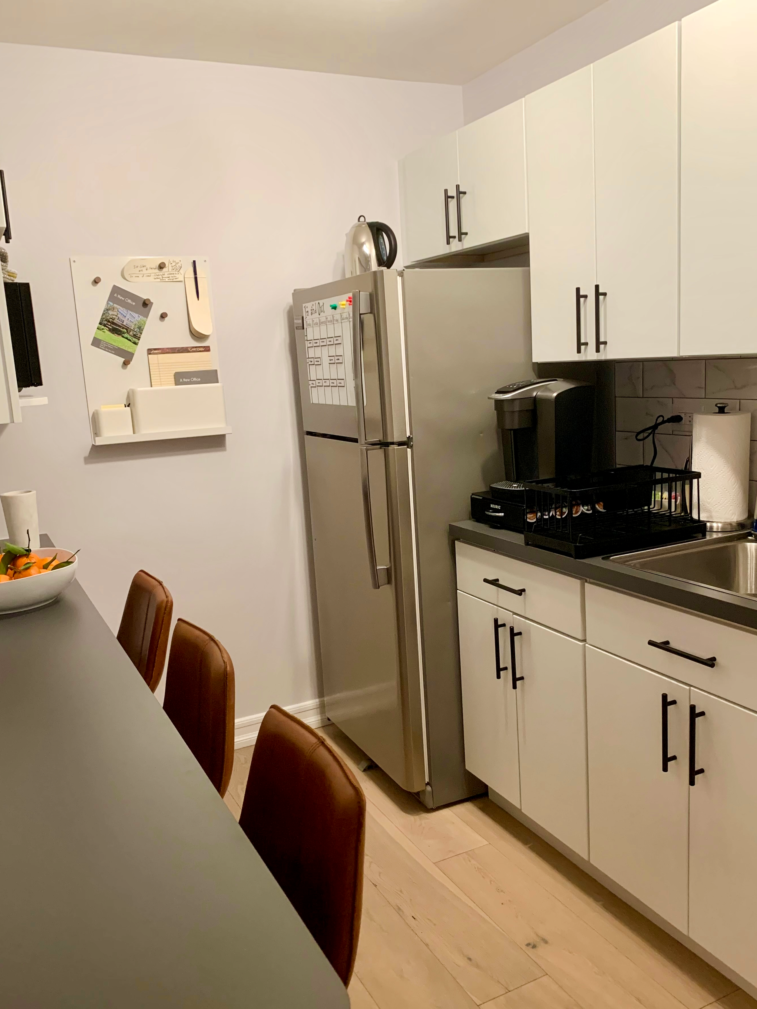 The staff  kitchen/lounge in a therapy office for rent in Brooklyn, with white cabinets, a refrigerator, a coffee maker; a counter with tan leather stools and a bowl of snacks.
