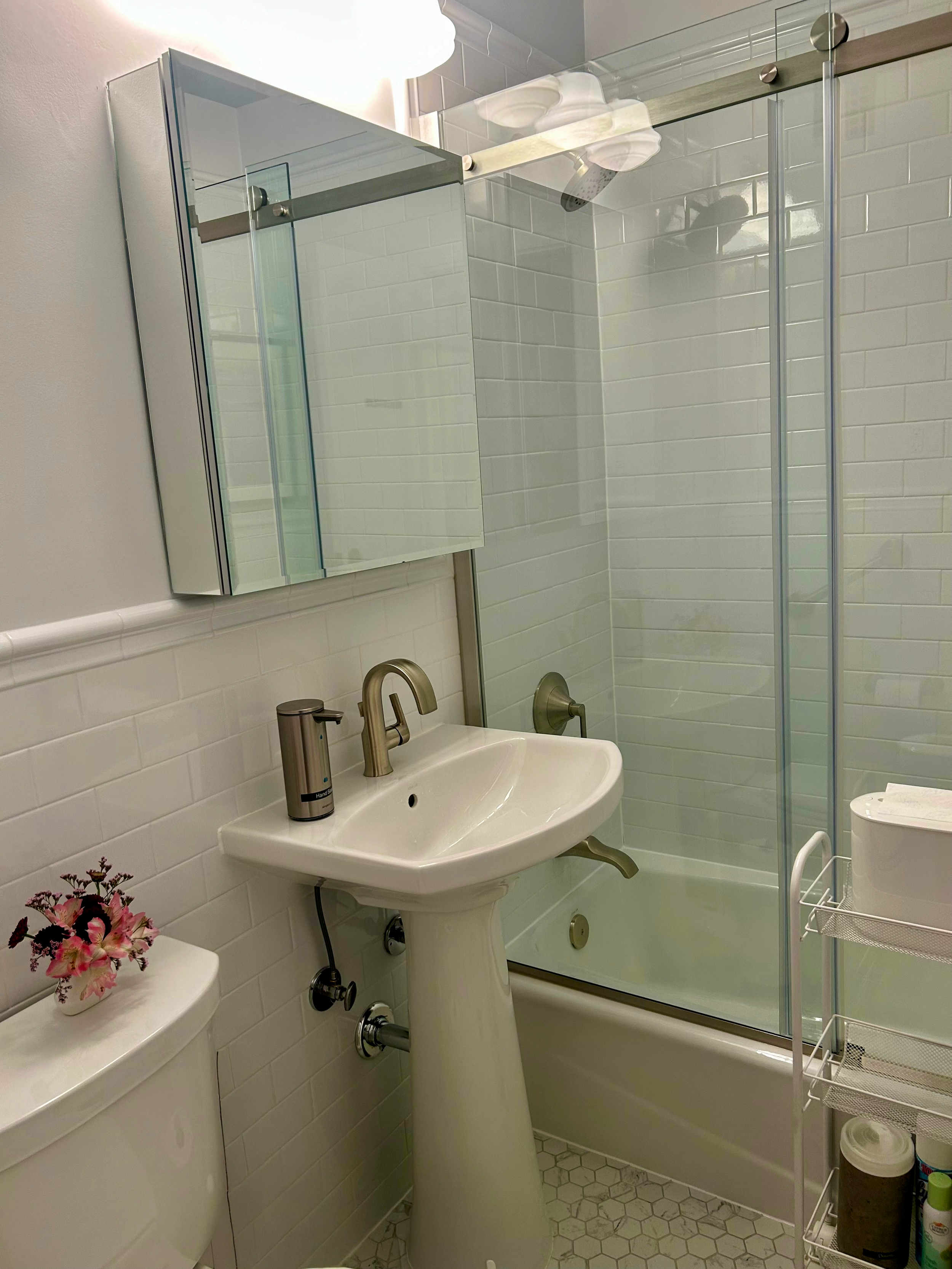 The bathroom of a therapy office for rent in Brooklyn, with a white pedestal sink, a mirror cabinet above the sink, a toilet with a vase of pink flowers nearby, and a glass-enclosed shower with white tiled walls.