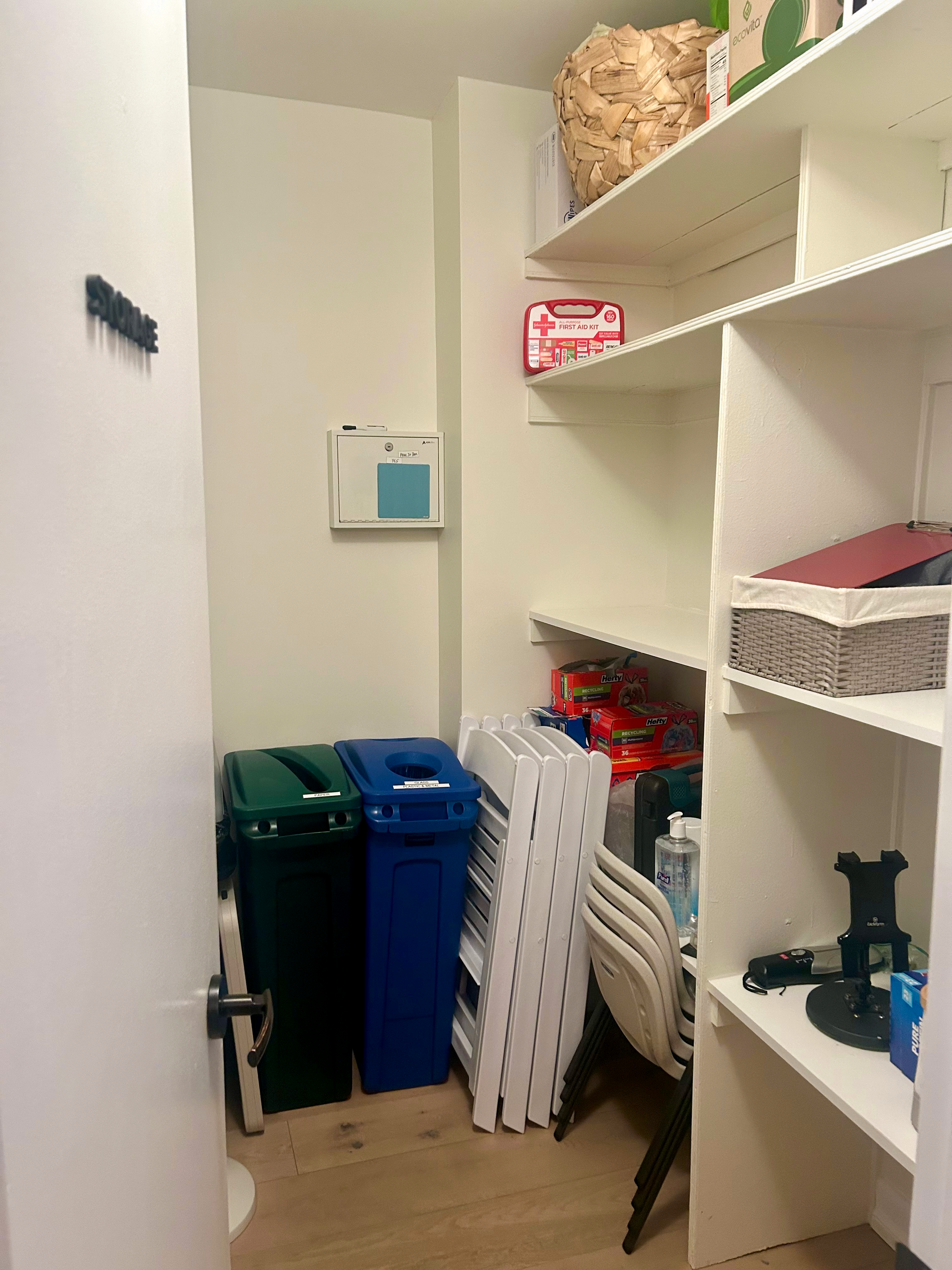 A storage closet in a therapy office for rent in Brooklyn, with white shelves containing various items, including a woven basket, first aid kit,, and cleaning supplies, with trash bins and folding chairs.